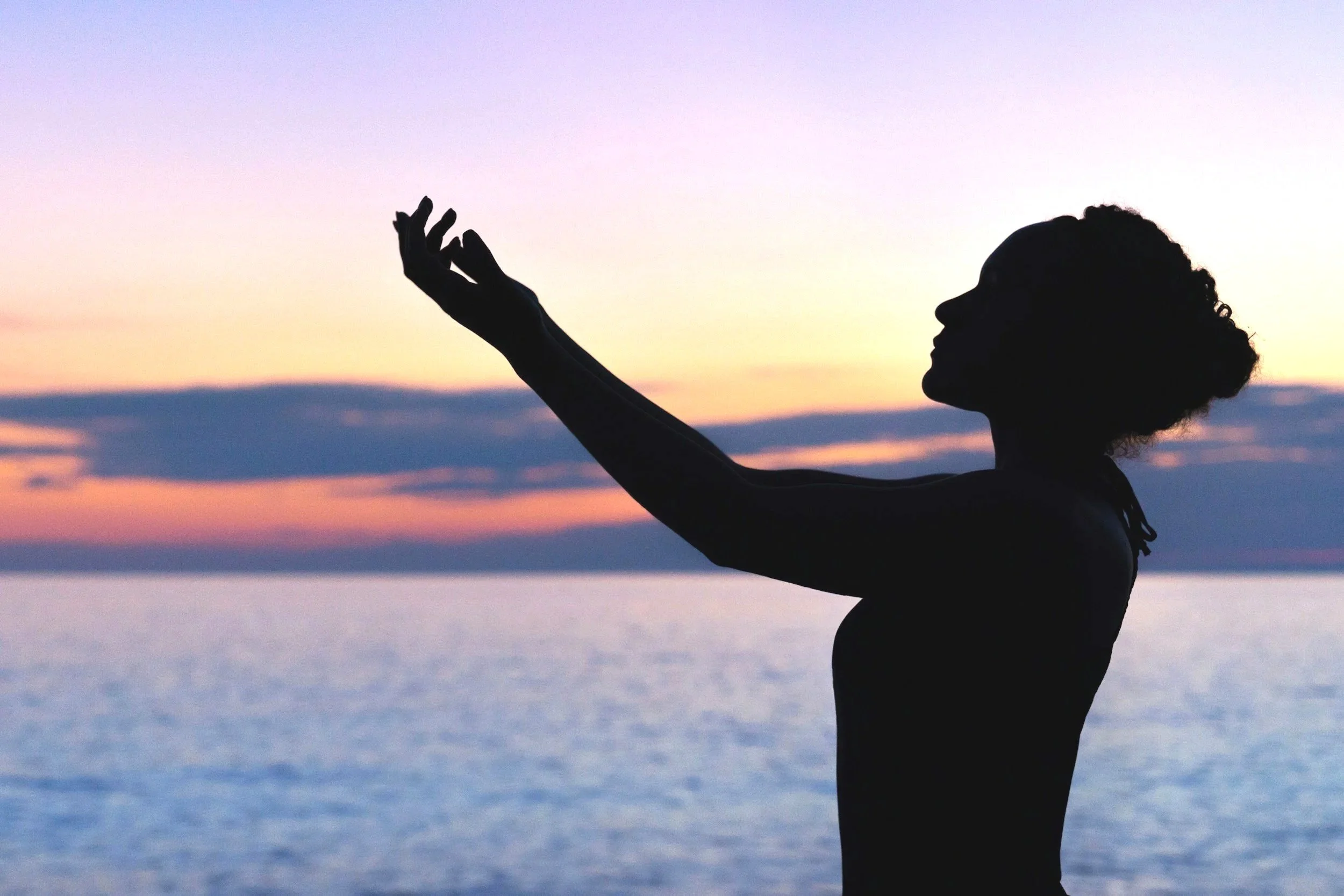 Silhouette of a woman with curly hair at sunset near the water, raising one arm with her hand in a graceful gesture.