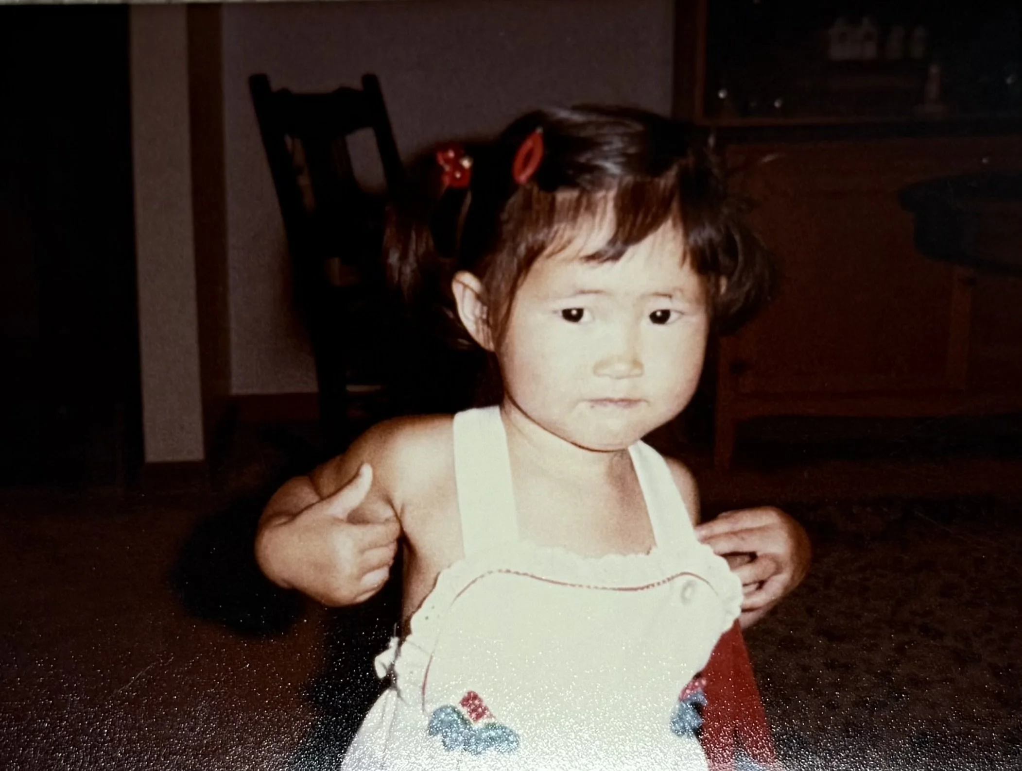 A young girl with short dark hair, wearing a white sleeveless top with embroidered designs, is sitting on the floor with her hands near her shoulders, in a room with wooden furniture and a carpet.