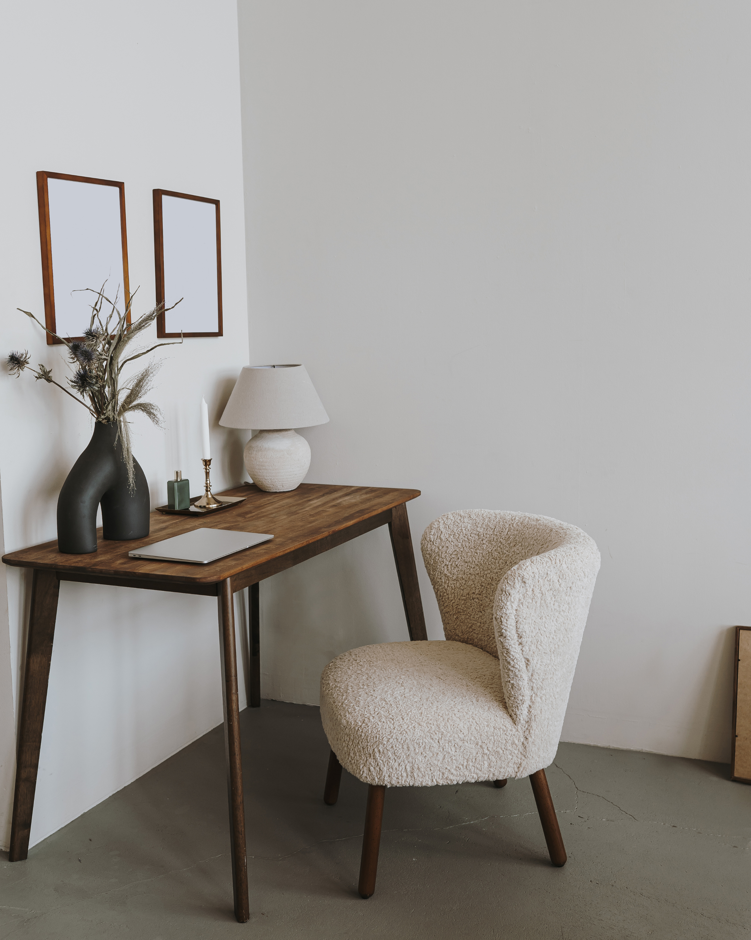 Un bureau en bois avec une lampe, un vase avec des plantes séchées, une bougie, et un ordinateur portable. À côté, une chaise en tissu beige.