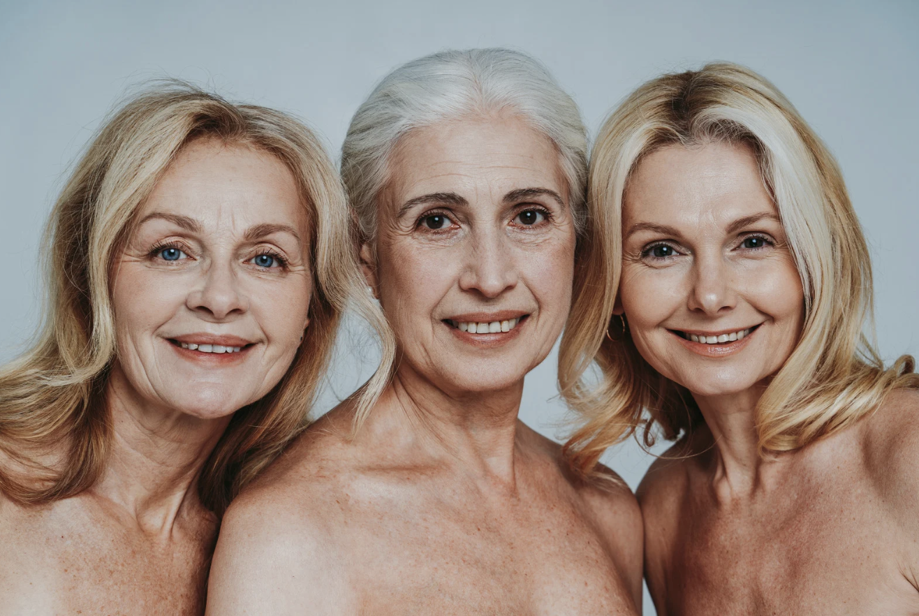 Trois femmes âgées souriantes, topless, debout ensemble contre un fond neutre.