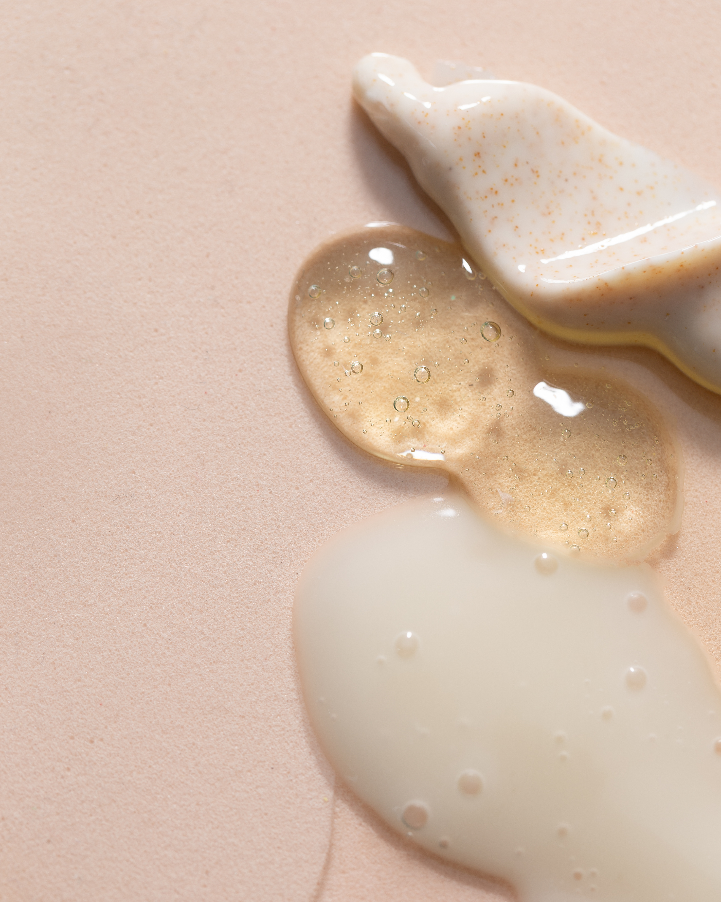 Gouttes de différents liquides sur une surface rose pâle, dont un blanc opaque, un beige translucide avec des bulles, et un liquide blanc mélangé à un liquide de couleur beige.