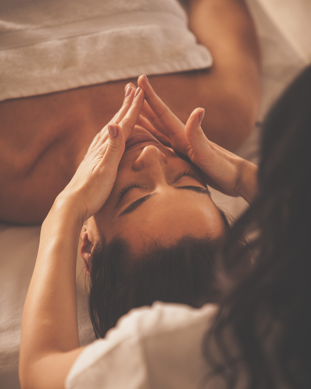 Une femme reçoit un massage du visage avec ses mains, allongée sur une table, ambiance chaleureuse et relaxante.