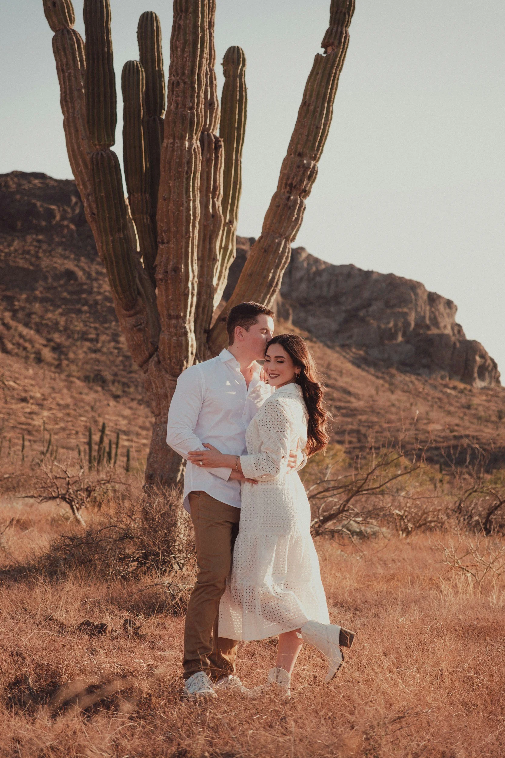 A couple stands close together outdoors in a desert landscape with a large cactus and rocky hills in the background, smiling and embracing.