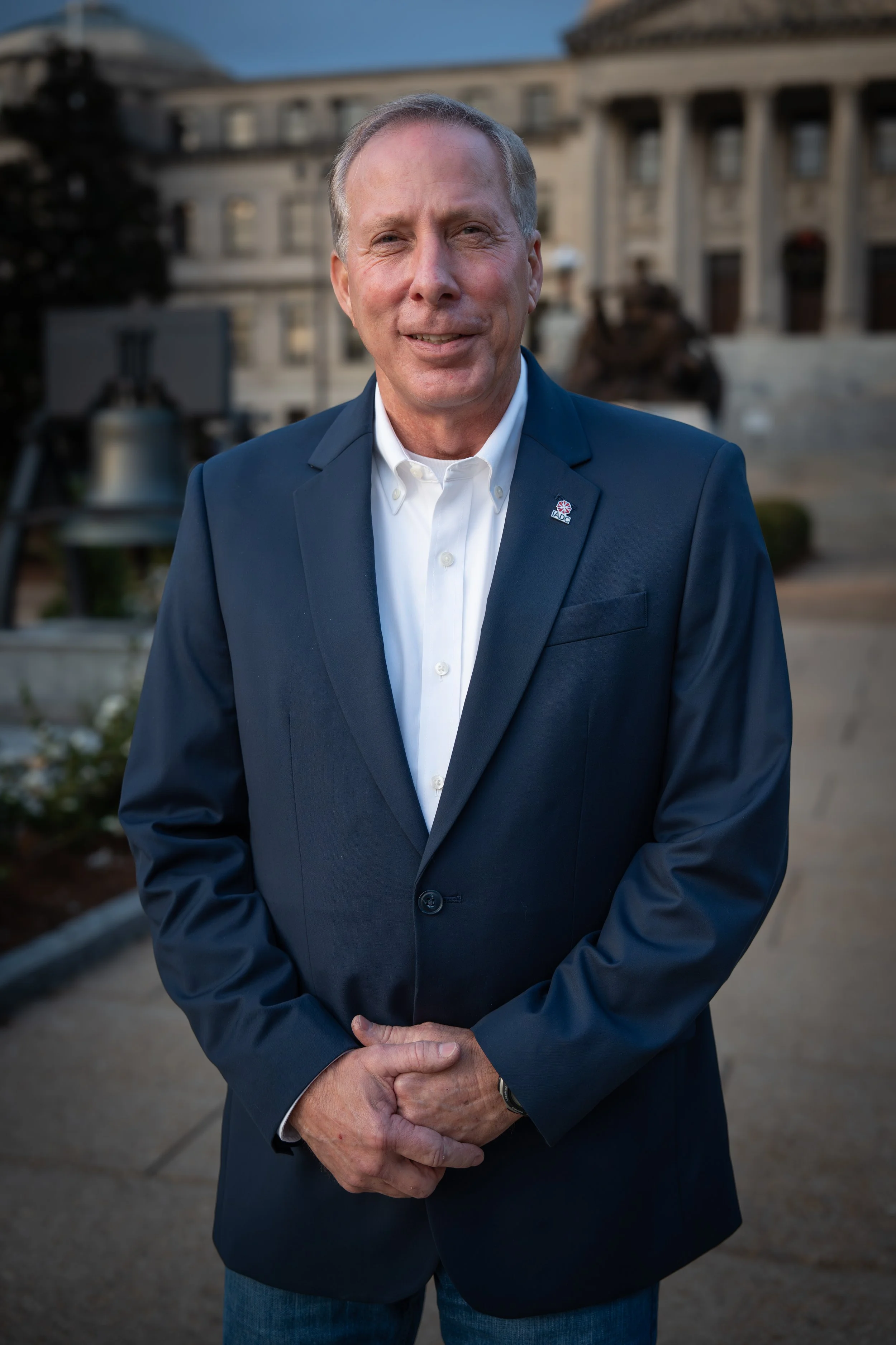 Man in a dark blue suit and white shirt standing outdoors in front of a historic building, smiling.