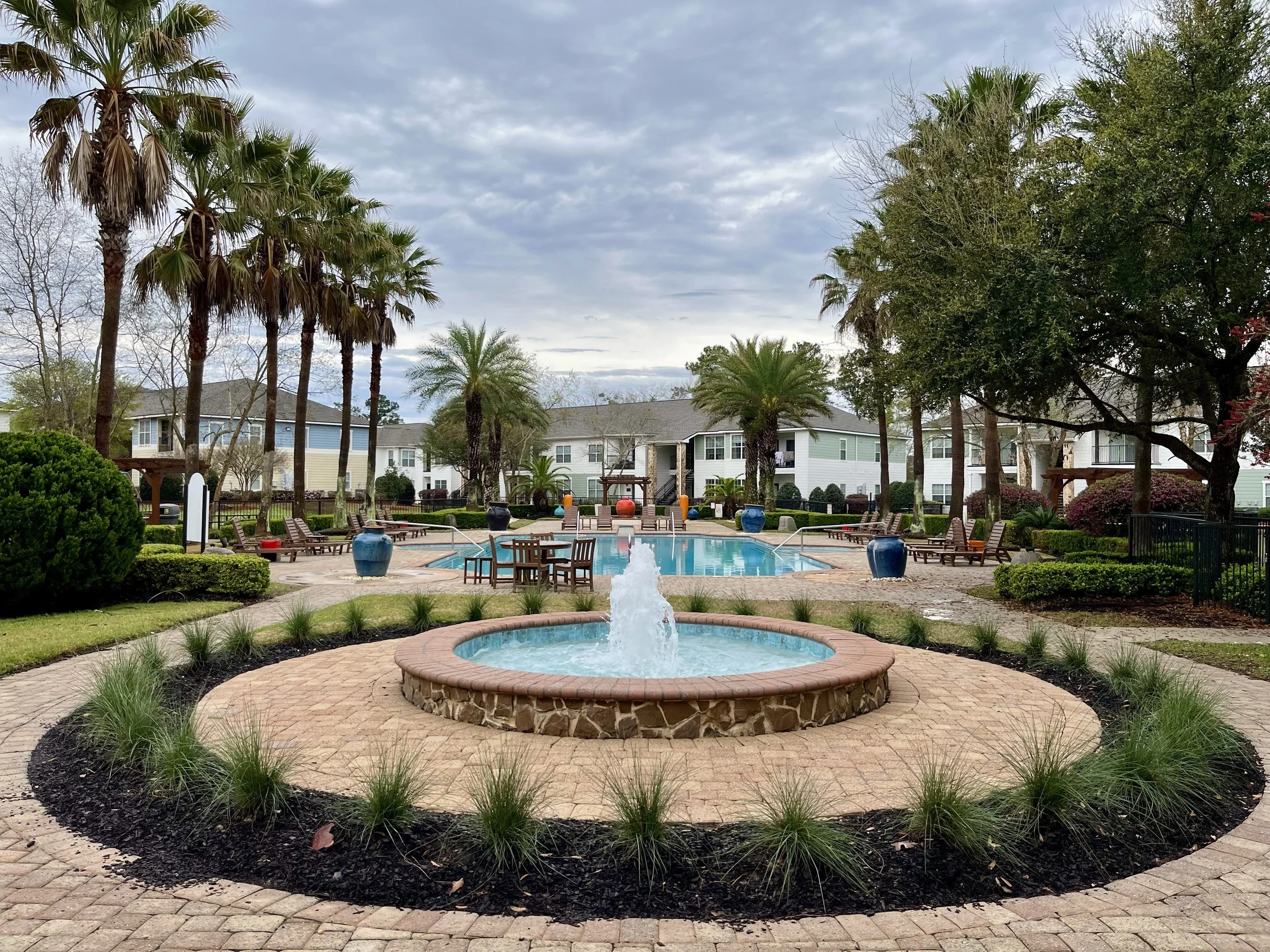 Community swimming pool area with palm trees, lounge chairs, a small fountain in the foreground, and residential buildings in the background under a cloudy sky. Villas Bon Secour
