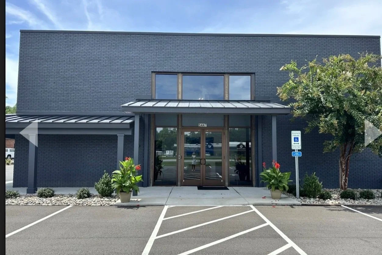 Modern commercial building with black brick exterior, glass entrance, and parking lot with designated handicapped parking spaces and signs. Potted plants and small shrubs decorate the entrance area.