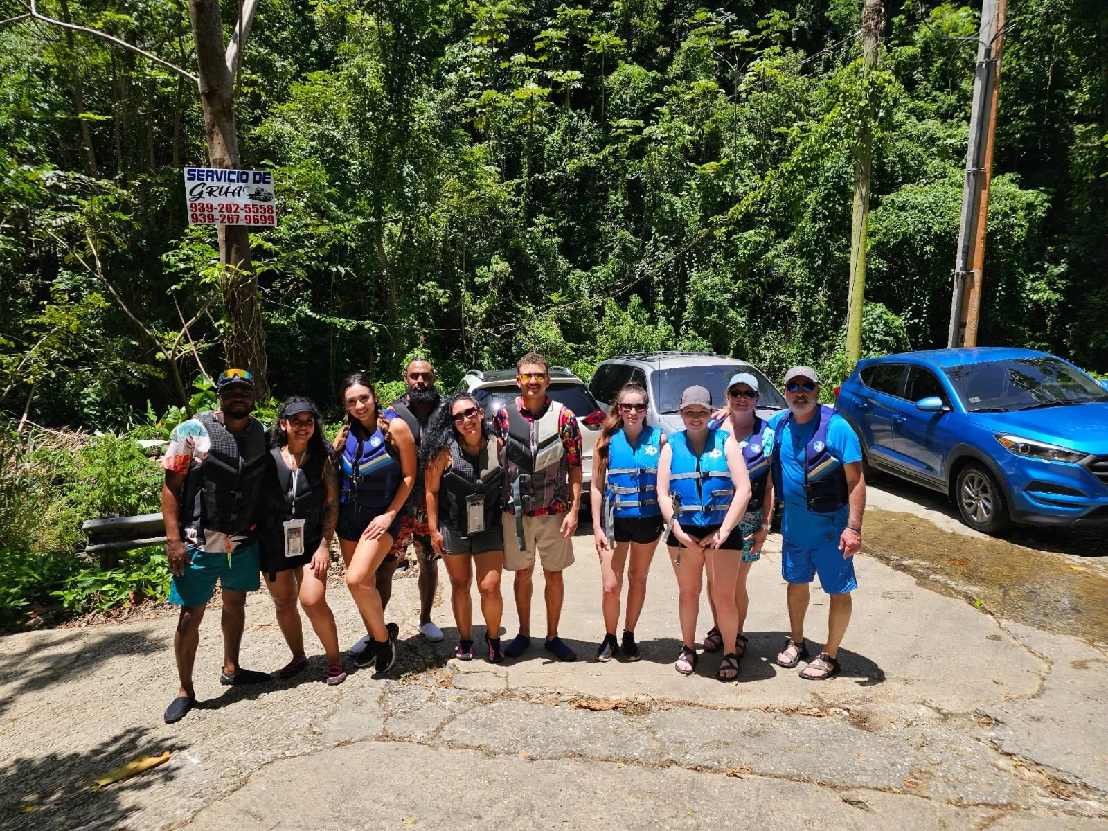 Group of ten people standing outdoors in front of cars and trees, wearing life vests and casual outdoor clothing, ready for a boat tour or adventure.