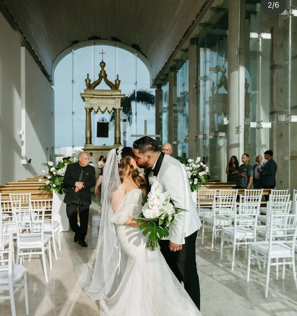 A bride and groom share their wedding kiss inside a modern church with glass walls and high arched ceiling. The bride wears a white wedding gown with a veil and holds a bouquet of white flowers. The groom wears a white tuxedo jacket with black pants. In the background, guests are seated and standing, with some dressed in formal attire. The church is decorated with white flowers and greenery.
