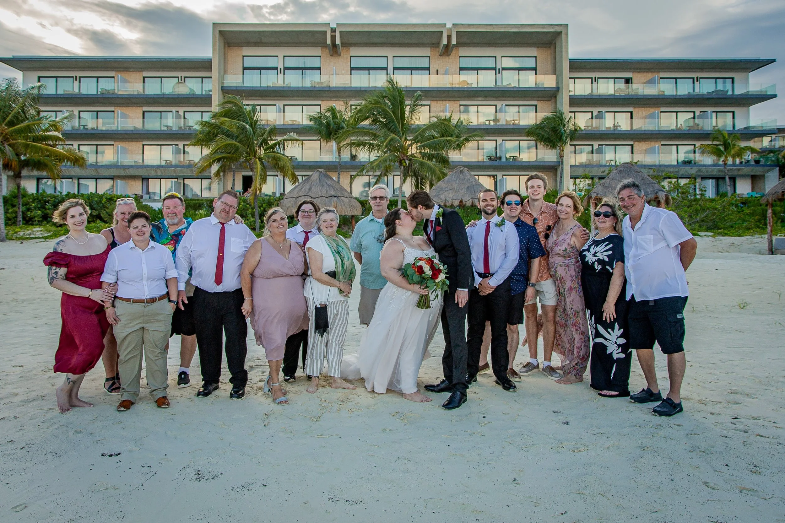 A group of people on a beach celebrating a wedding, with a modern hotel and palm trees in the background.