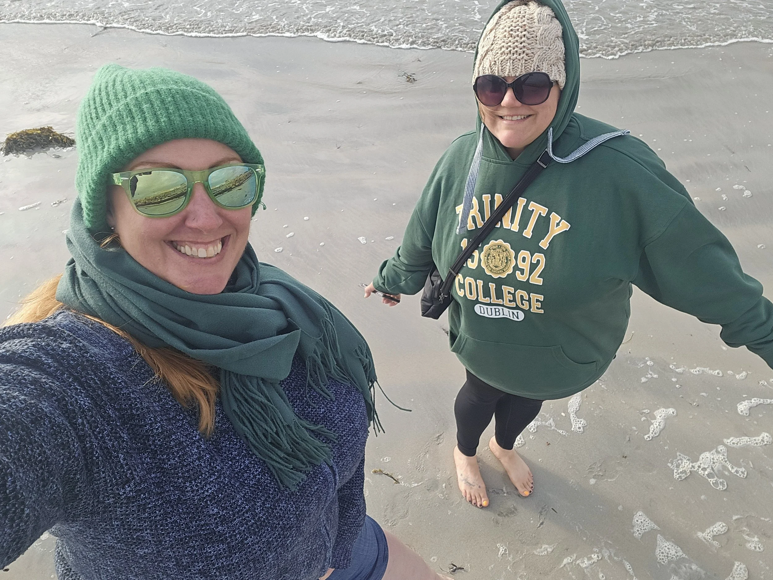 Two women taking a selfie on a beach with footprints in the sand, both smiling and wearing sunglasses and warm clothing.