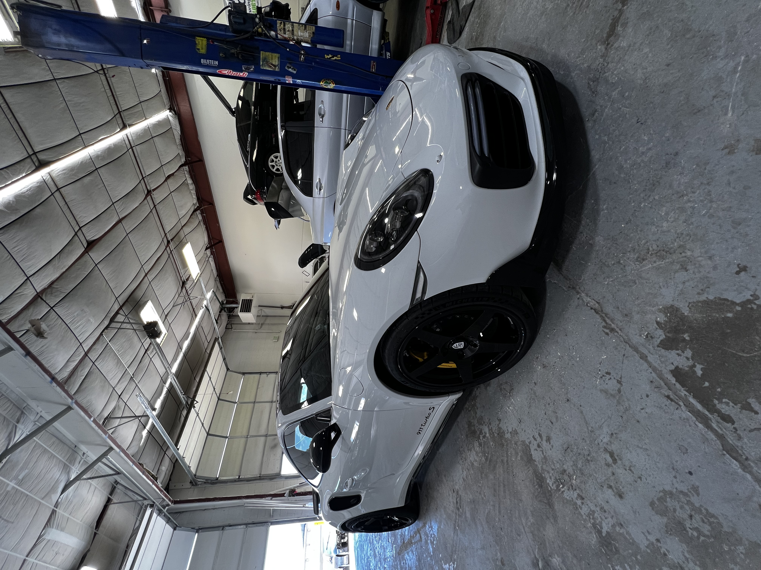 A white Porsche with black wheels parked inside a garage. Other cars are also visible nearby, one of which is suspended on a hydraulic lift. The garage has a high ceiling with insulation and fluorescent lighting.