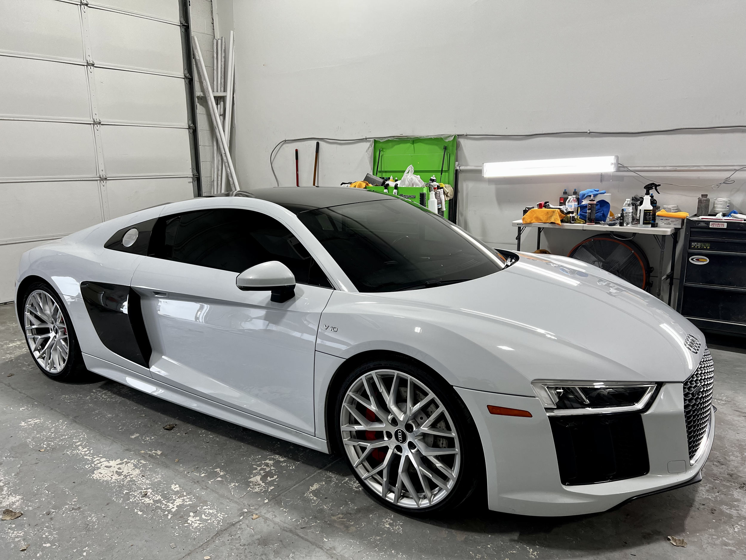 A white Audi R8 parked inside a garage. The garage has some tools and supplies on a workbench and a tool chest in the background.