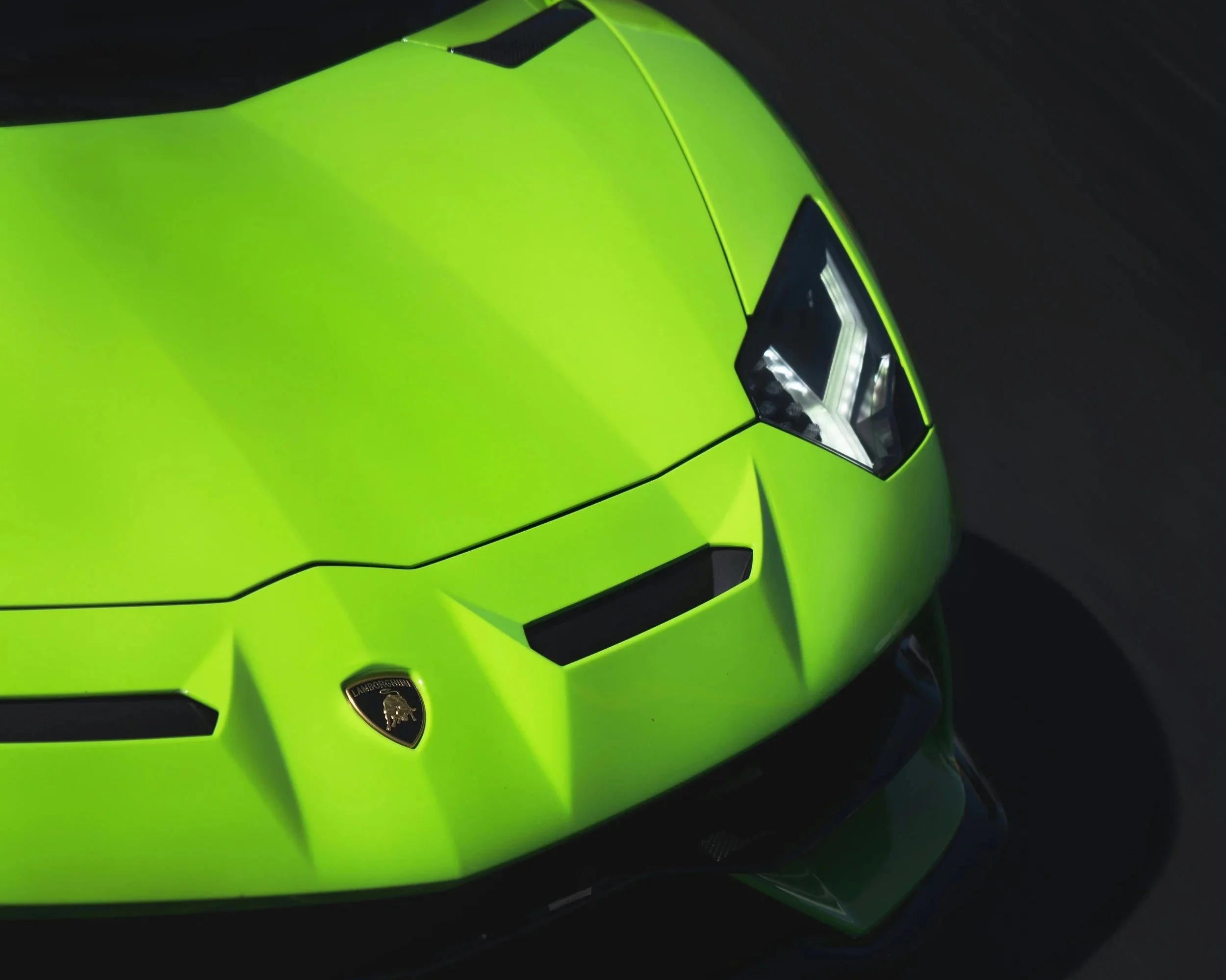 Close-up of a bright green Lamborghini sports car with black accents, featuring the Lamborghini crest on the front hood and a distinctive angular headlight design.