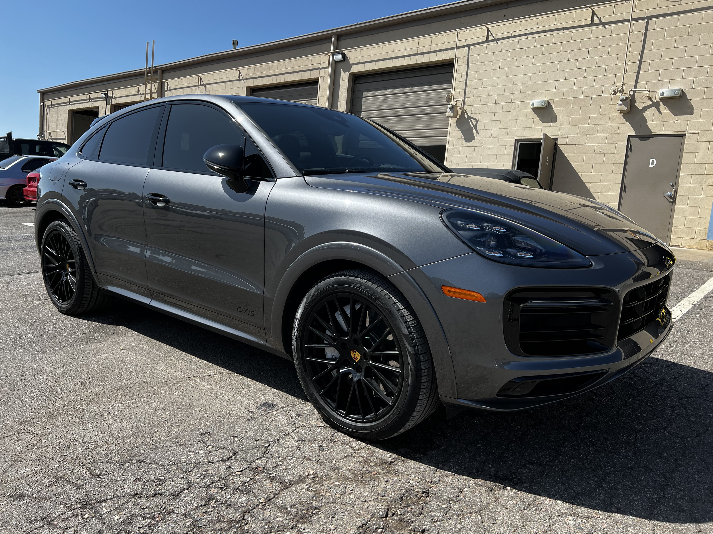 Gray Porsche Macan GTS parked outdoors in a lot with some other cars and a beige industrial building in the background.