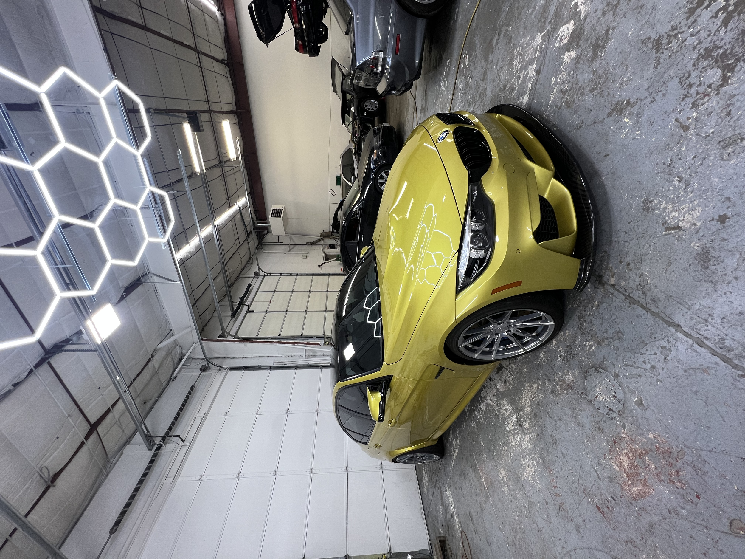 A yellow sports car parked inside a garage with other cars in the background. The garage has a high ceiling with hexagon-shaped LED lights.