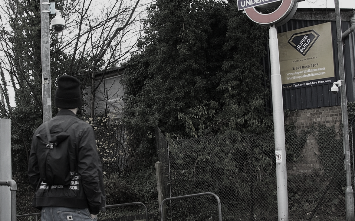 A person wearing a black beanie, jacket, and backpack standing near a street sign for the London Underground in an urban setting with trees and a fence.