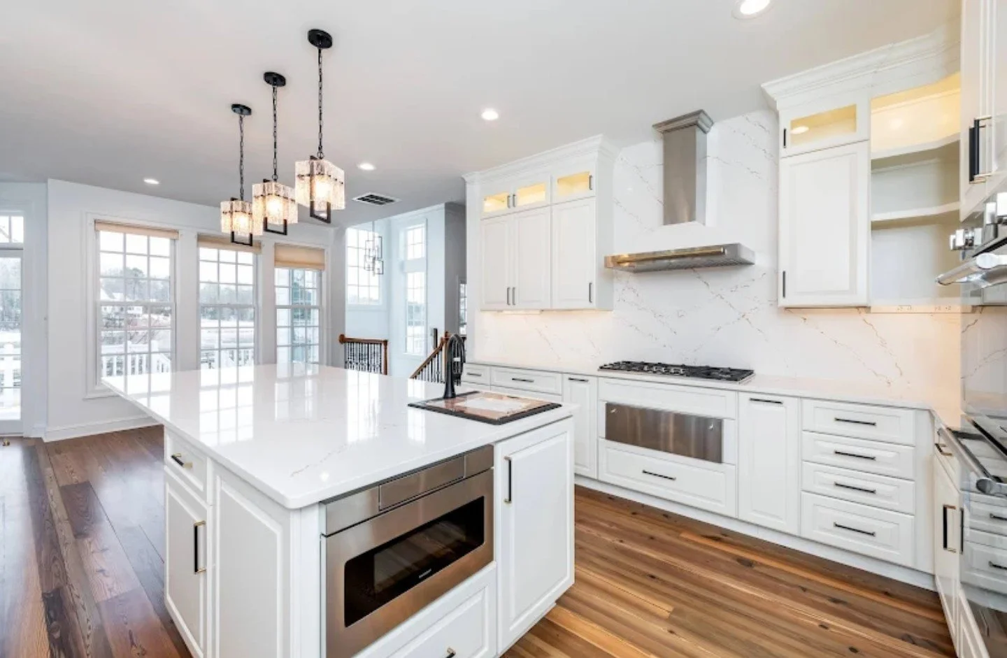 Jewel Box Homes - Fords Colony Williamsburg, VA Bright modern kitchen with white cabinets, marble backsplash, and wooden floors. Kitchen island with built-in microwave, pendant lights overhead, large windows bringing natural light. 