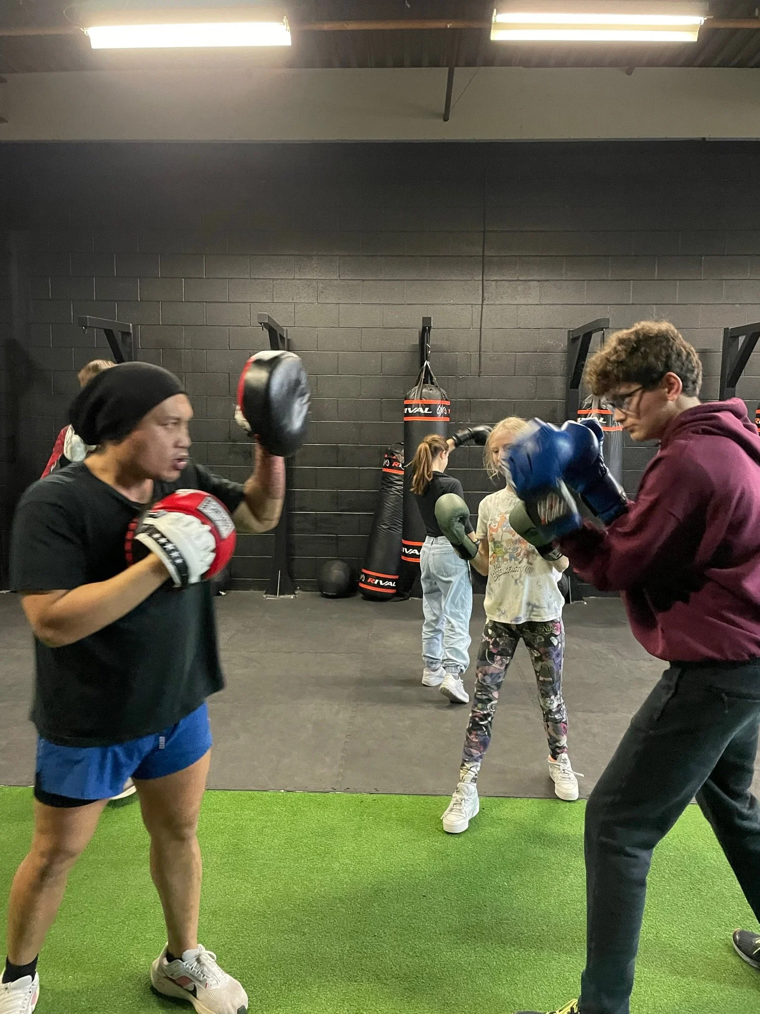 People practicing boxing in a gym with punching bags and children in the background.