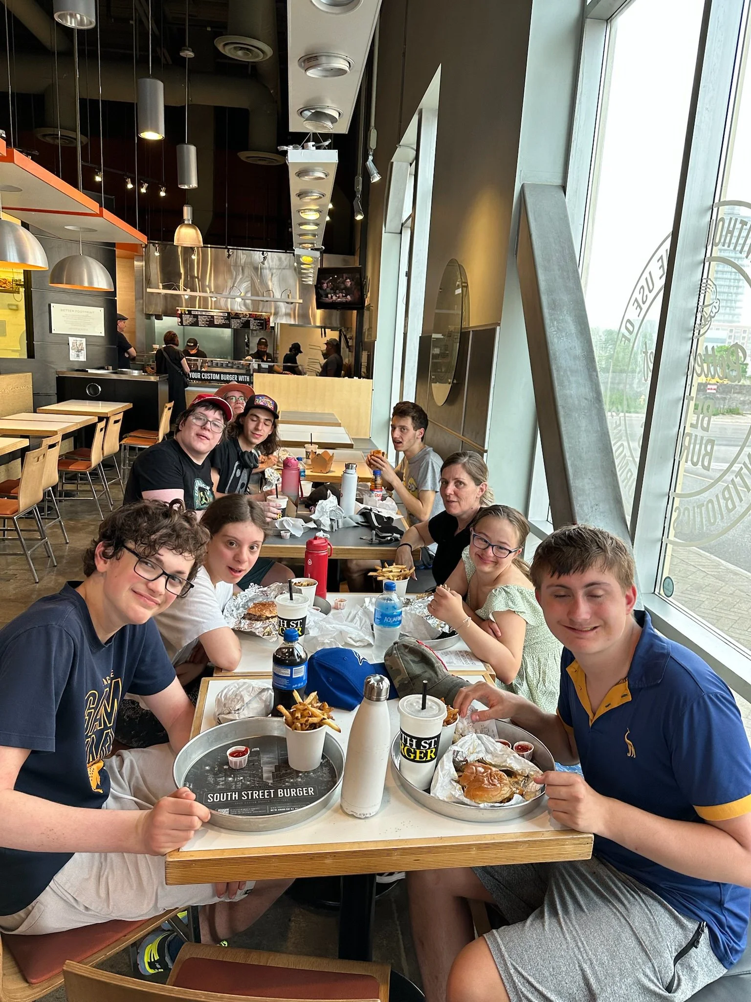 A group of eight young people sitting at a table in a burger restaurant, enjoying burgers and fries, with some drinks on the table, inside a modern restaurant with large windows.