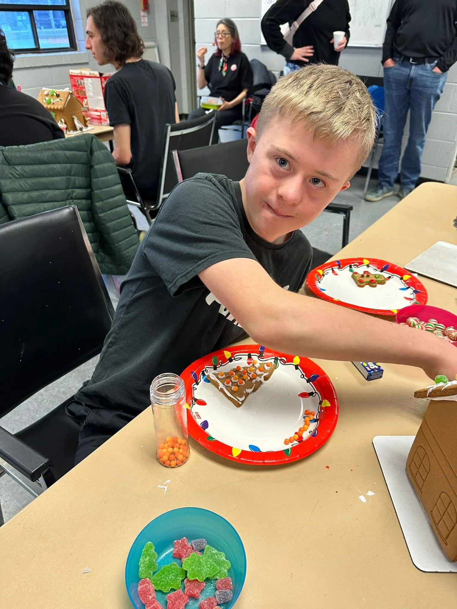 A young boy with blond hair wearing a black t-shirt, sitting at a table with decorated gingerbread cookies and colorful candies for a holiday celebration. Other people are in the background, some standing and some seated, in a room decorated for the 