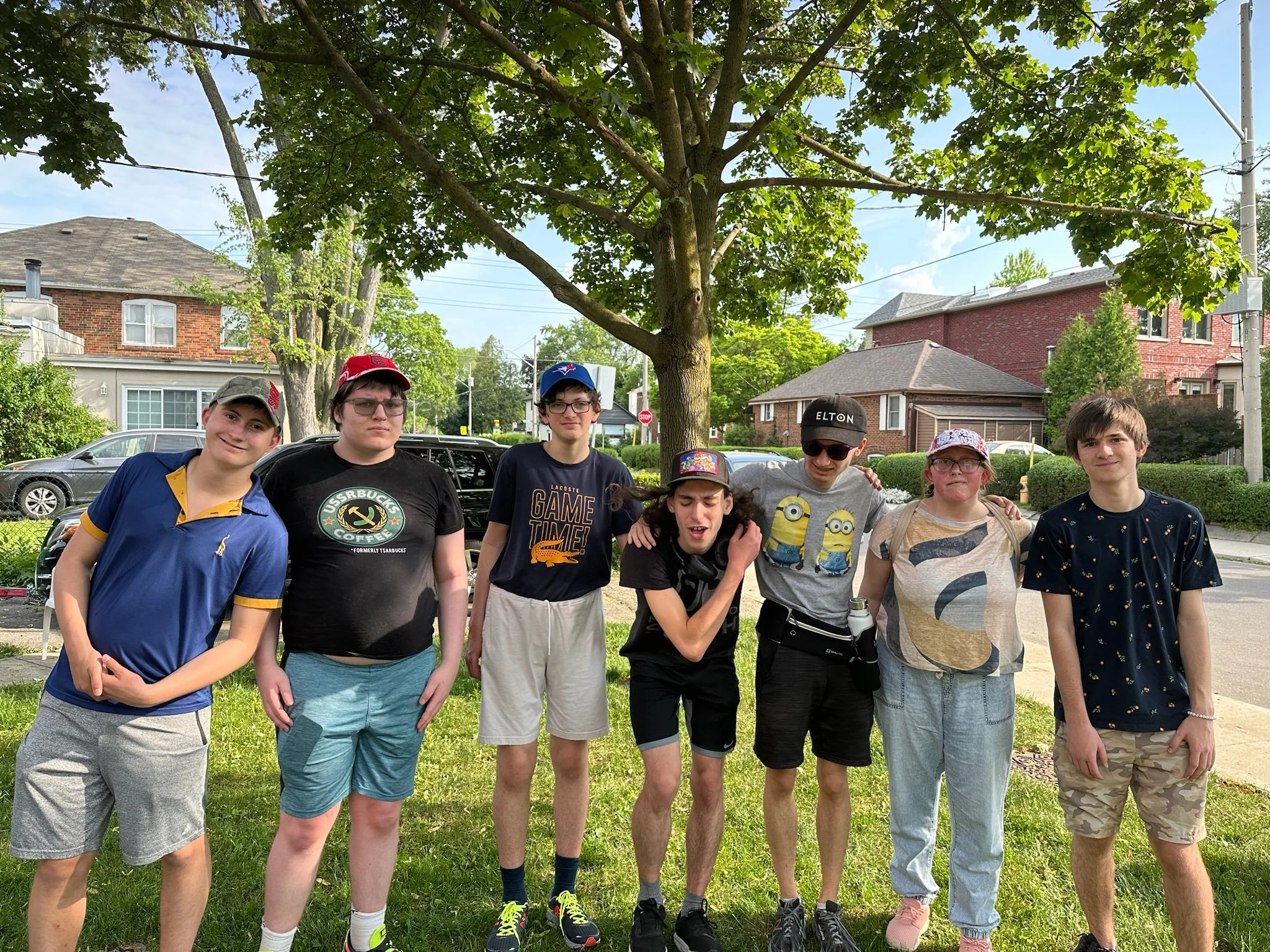 A group of eight young boys standing outdoors on a grassy area under a large tree, with residential houses and parked cars in the background. They are dressed casually, some wearing hats and glasses, and are posing for a photo.