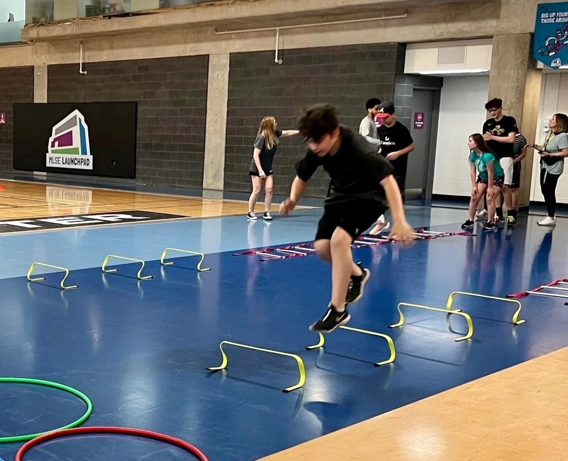 A boy practicing agility drills on a blue indoor gym floor, jumping over small hurdles, with other children and adults waiting in line behind him.
