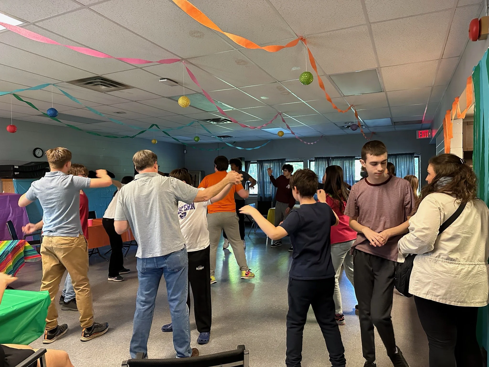 Teenagers and adults dancing at a party or social gathering in a decorated room with colorful streamers and hanging paper balls.