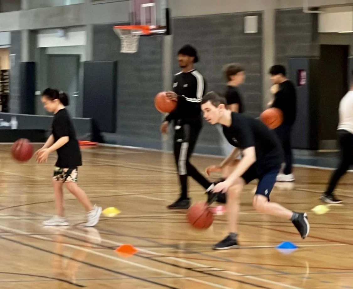 Children practicing basketball drills in a gym with a coach holding a basketball, cones on the floor, and a basketball hoop in the background.