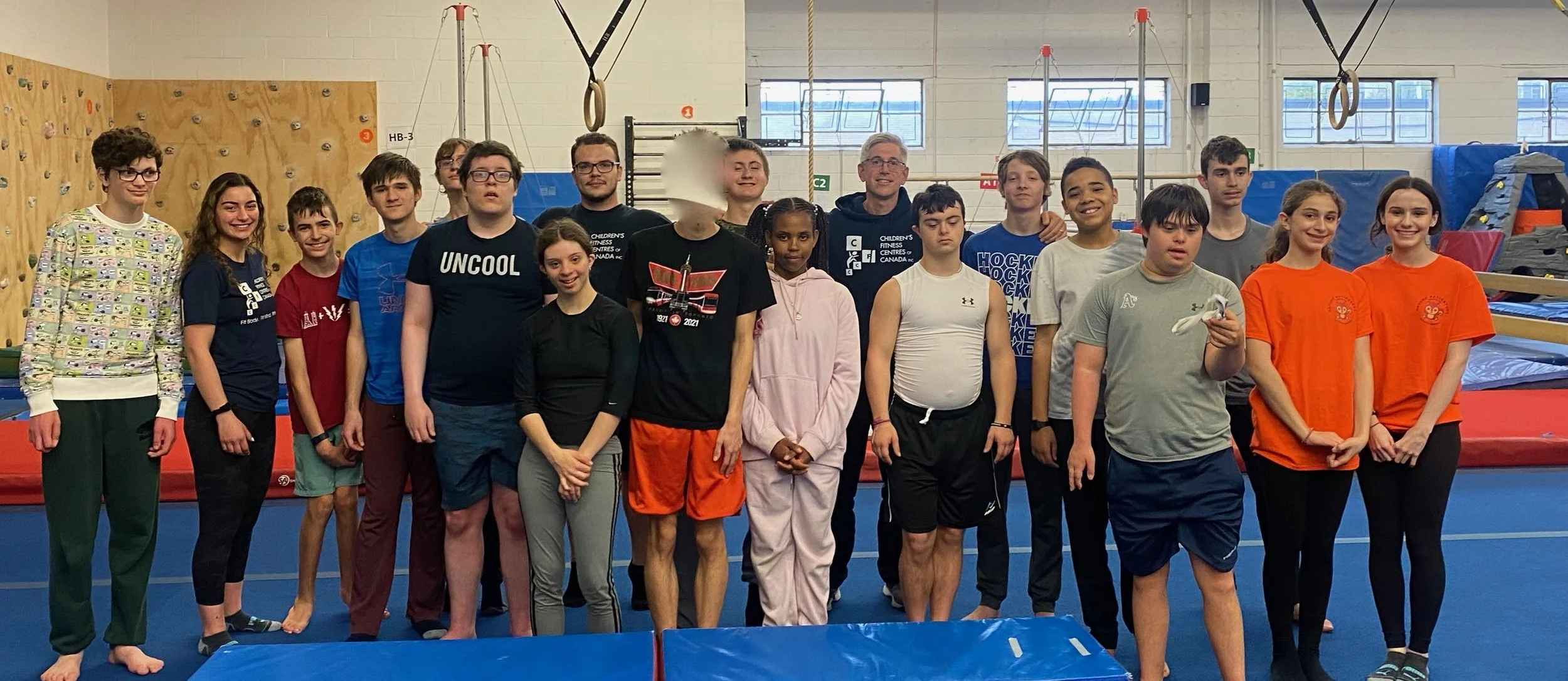 Group of young gymnasts and coaches standing on an indoor gymnastics facility floor, with equipment and climbing wall in the background.