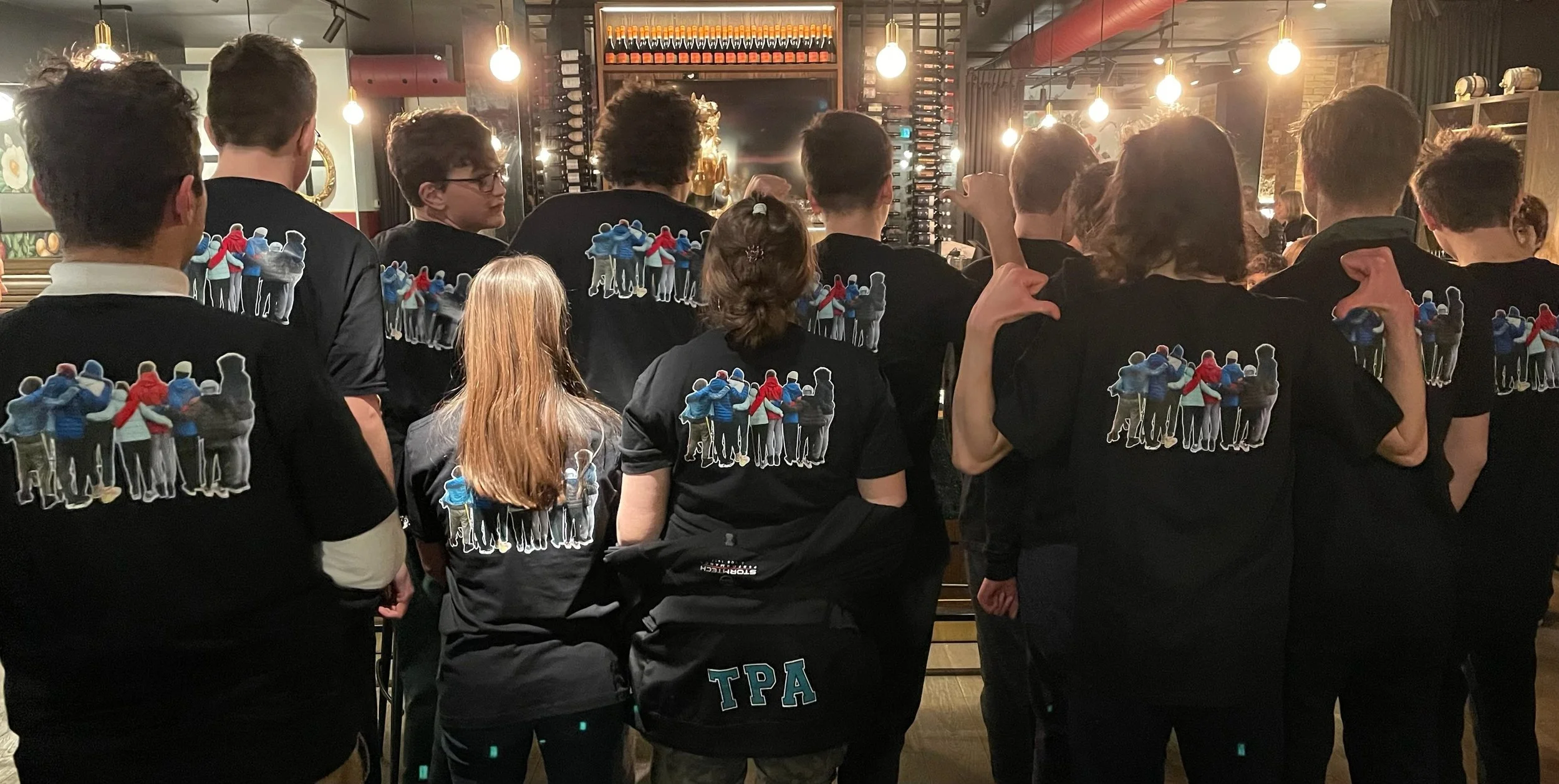 A group of people wearing black shirts with a graphic of a group hug printed on the back, gathered in a restaurant or bar with warm lighting and wine racks in the background.