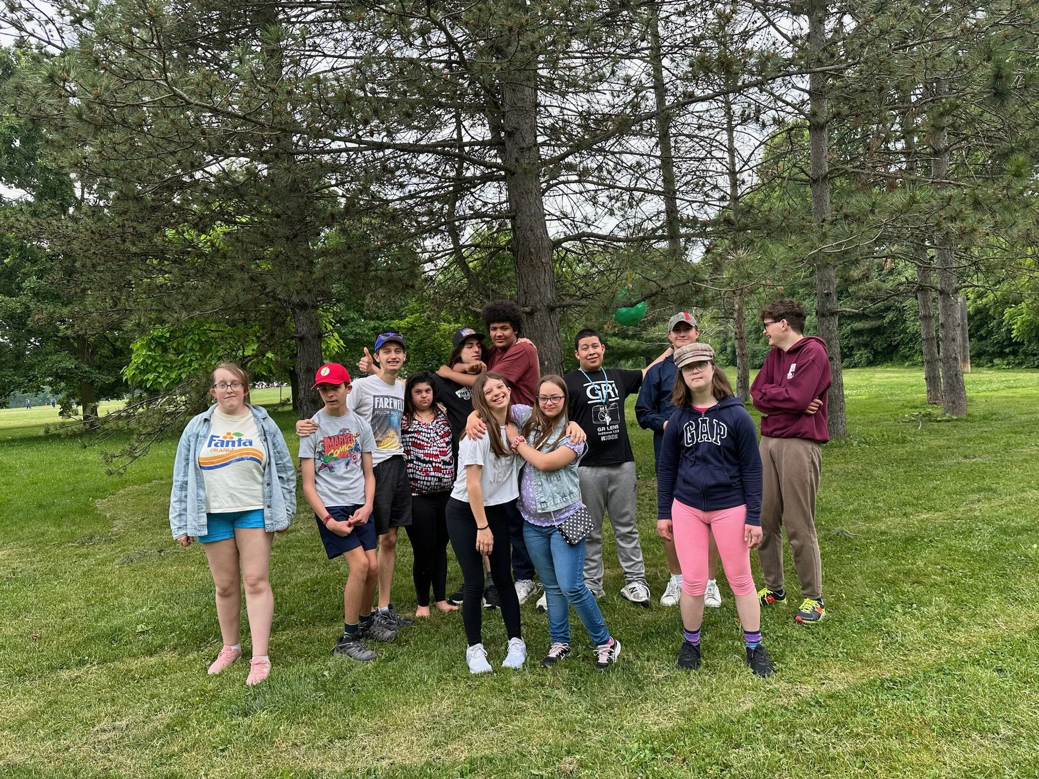 A group of people, mostly children and teenagers, gathered outdoors in a park-like setting with green grass and trees in the background, posing for a photo.