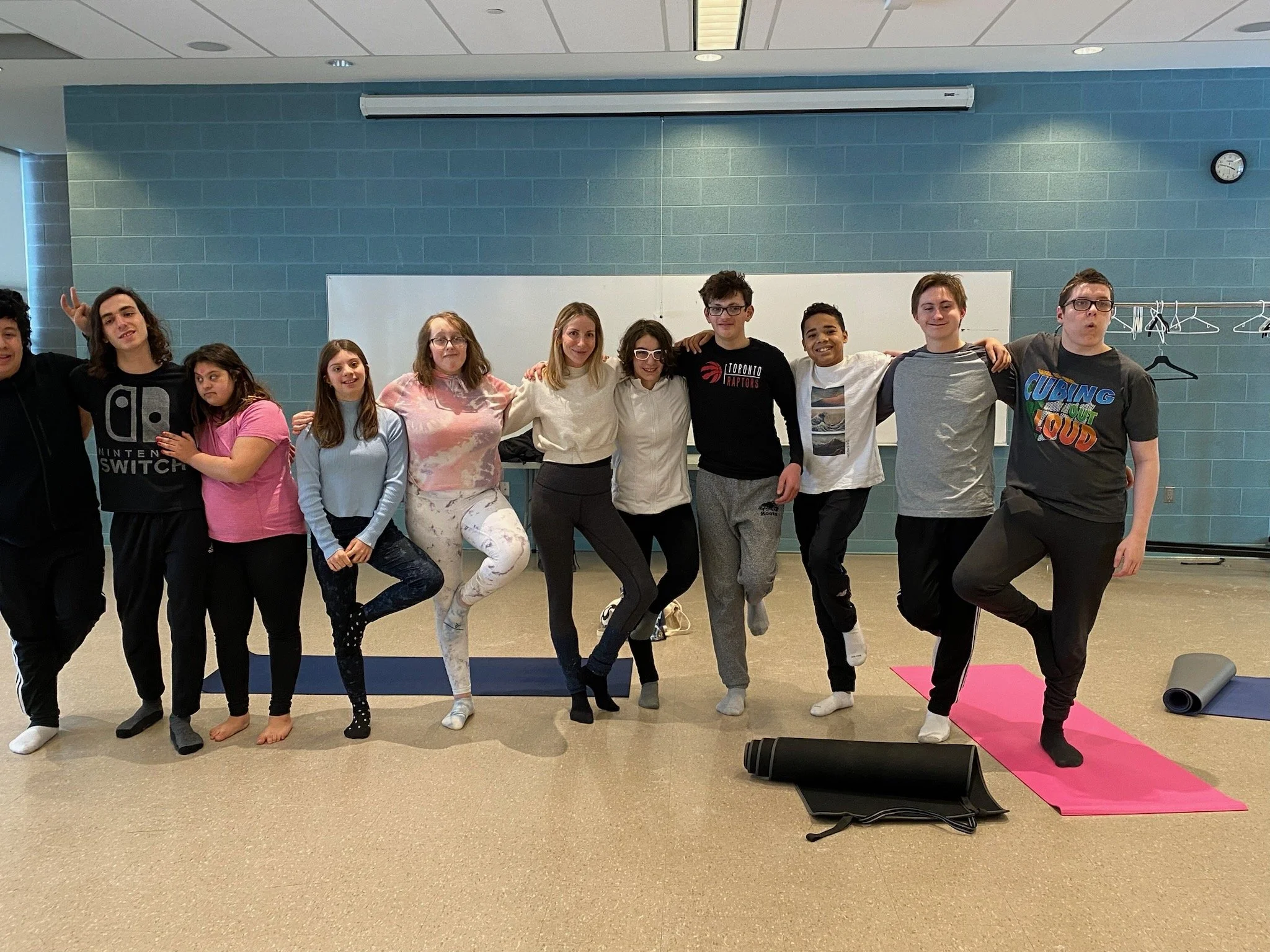 Group of eleven teenagers standing in a row indoors, some with arms around each other, at a yoga or dance studio, with yoga mats on the floor and a whiteboard and clock on the wall behind them.