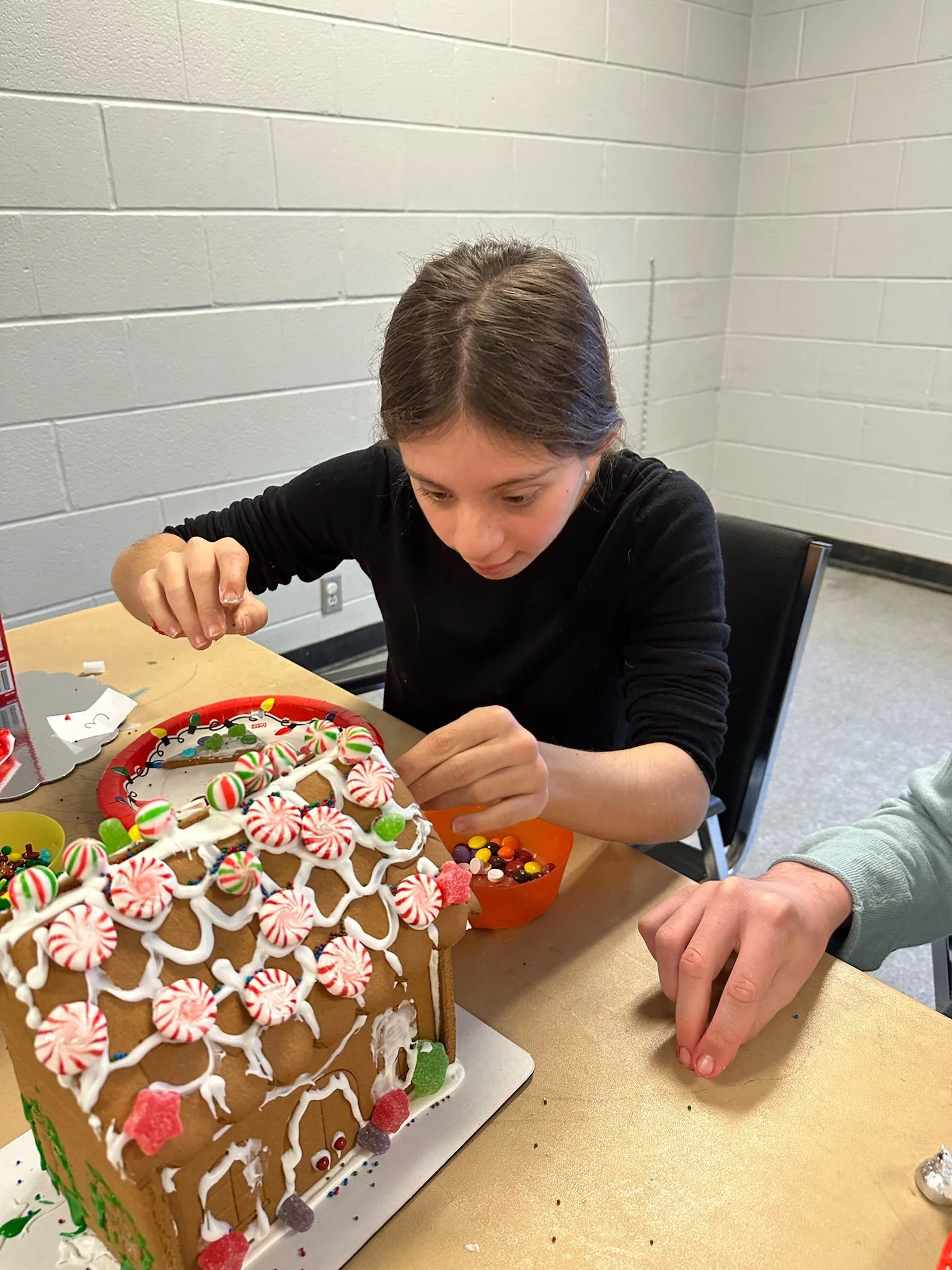 A young girl with brown hair decorating a gingerbread house with icing and candies.