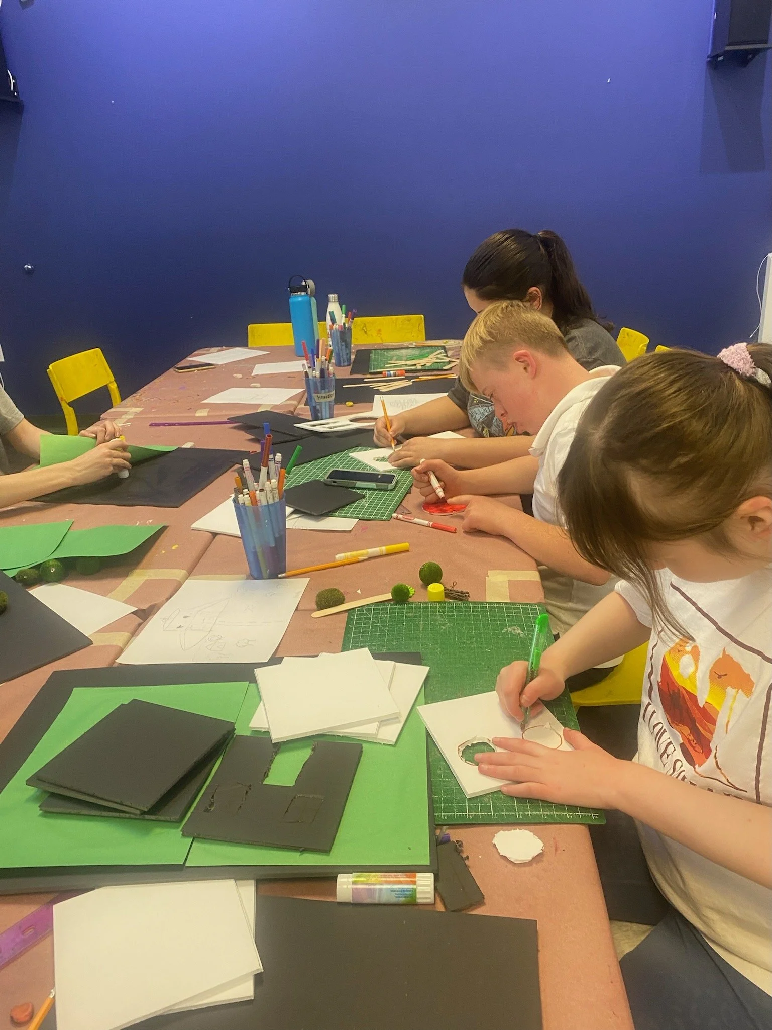Children engaging in arts and crafts at a table filled with paper, scissors, markers, and colored foam sheets.