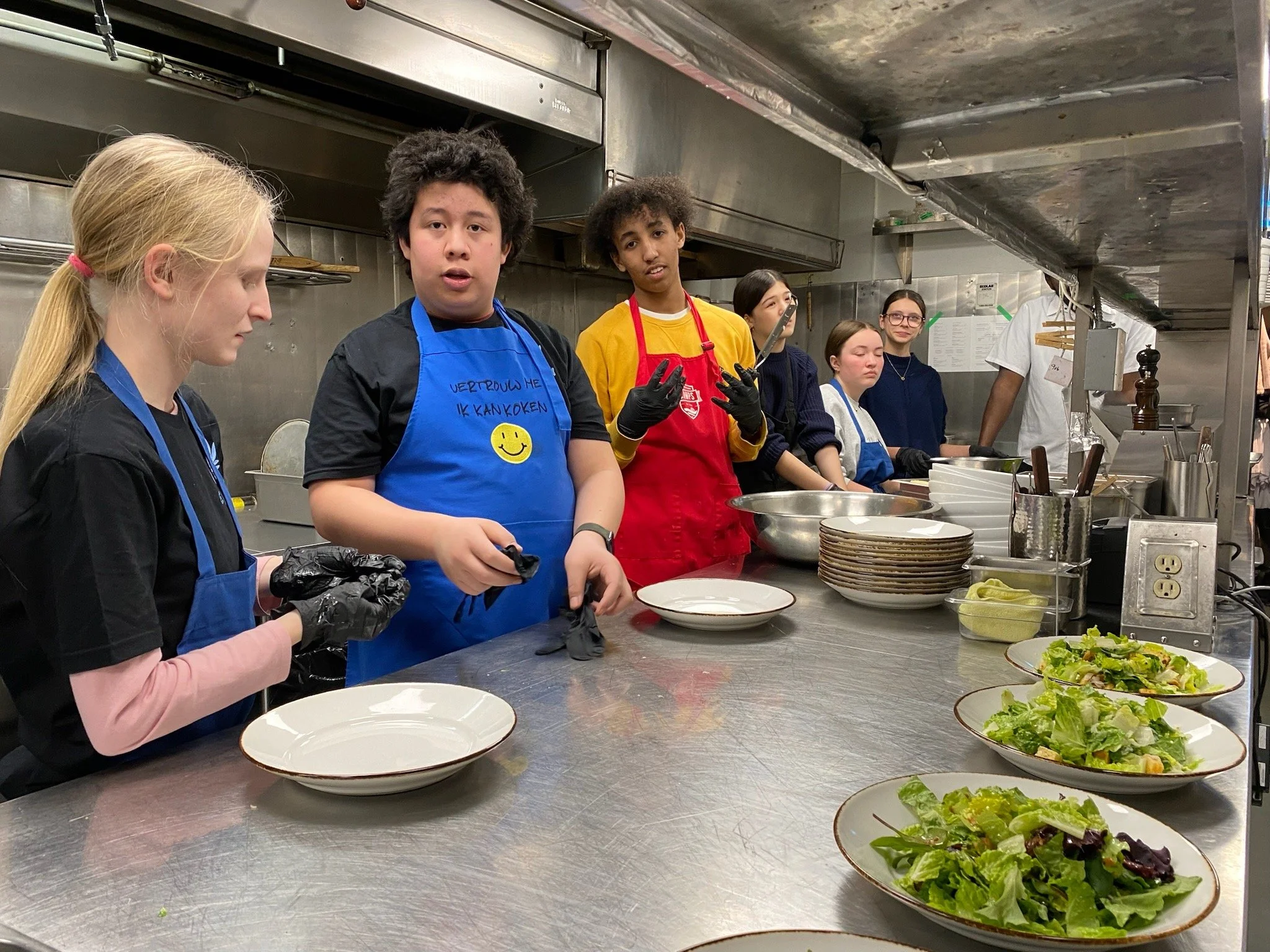 Group of children in a restaurant kitchen preparing salads, some wearing aprons and gloves, with plates of salad on the counter.