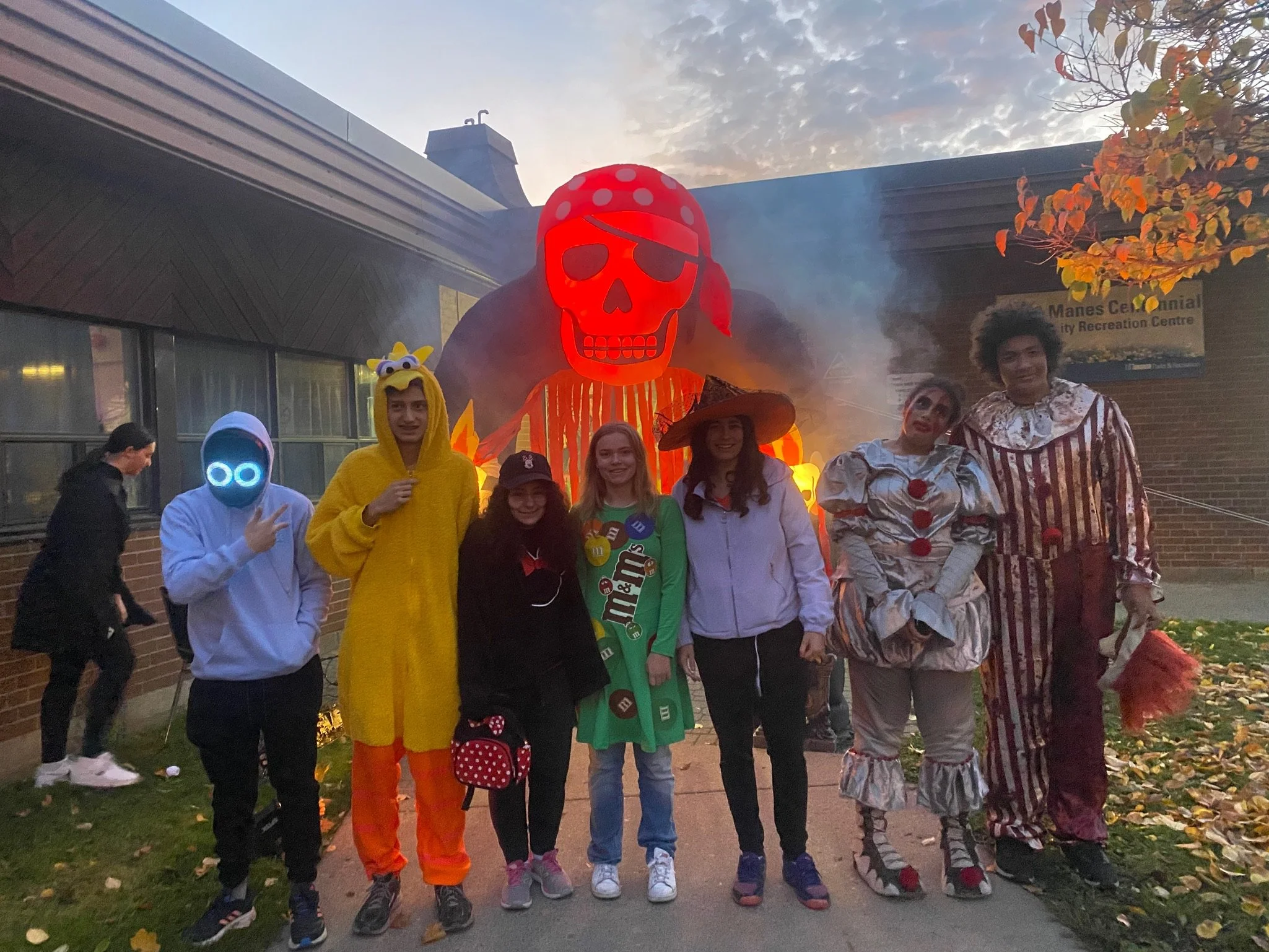 Group of people dressed in costumes standing outdoors in front of a large, illuminated, ghostly clown figure with a skull face at a Halloween or themed event.