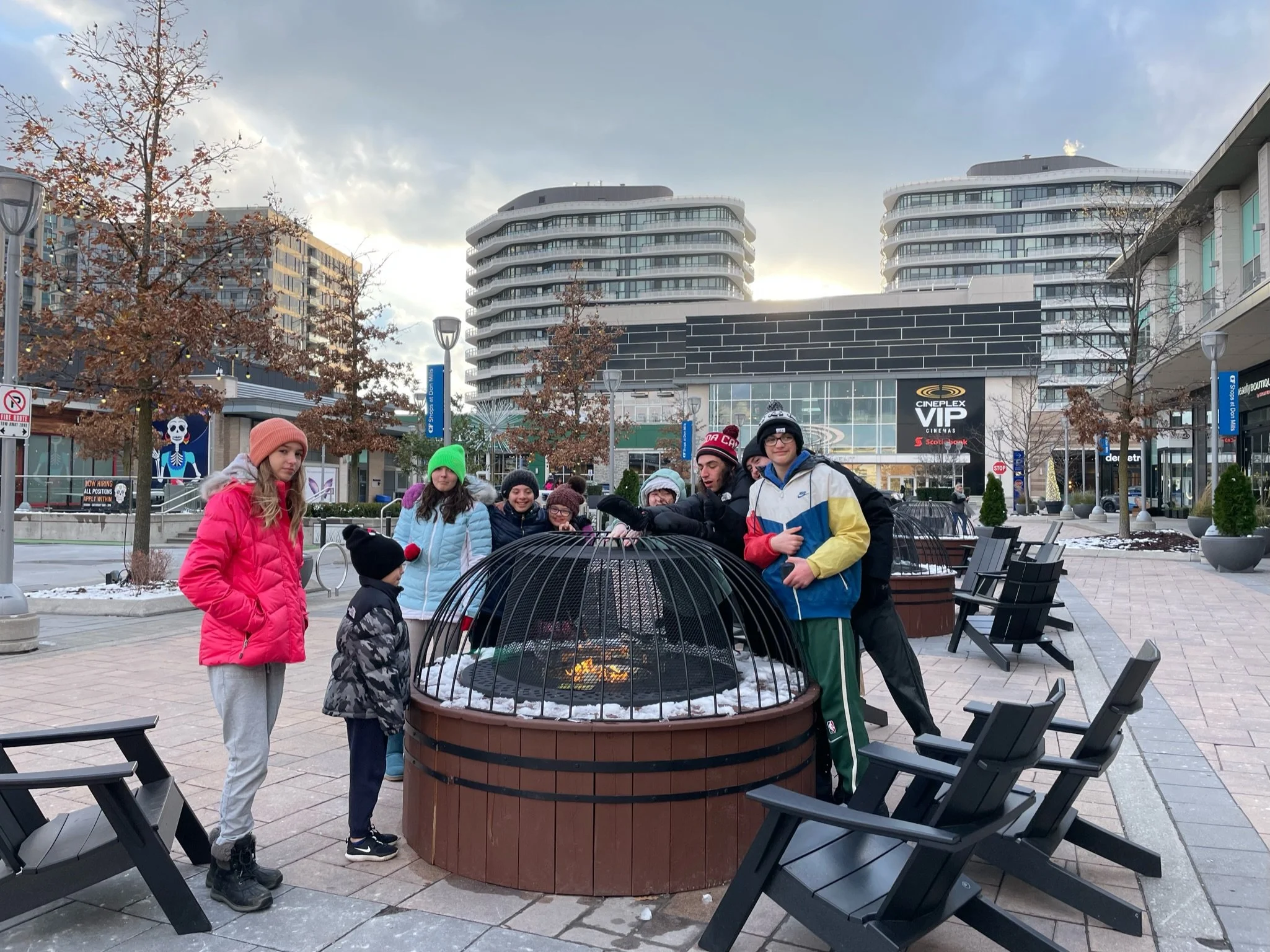 Group of people around a fire pit with a wire cage cover in an outdoor mall with modern buildings in the background, some wearing winter clothing.