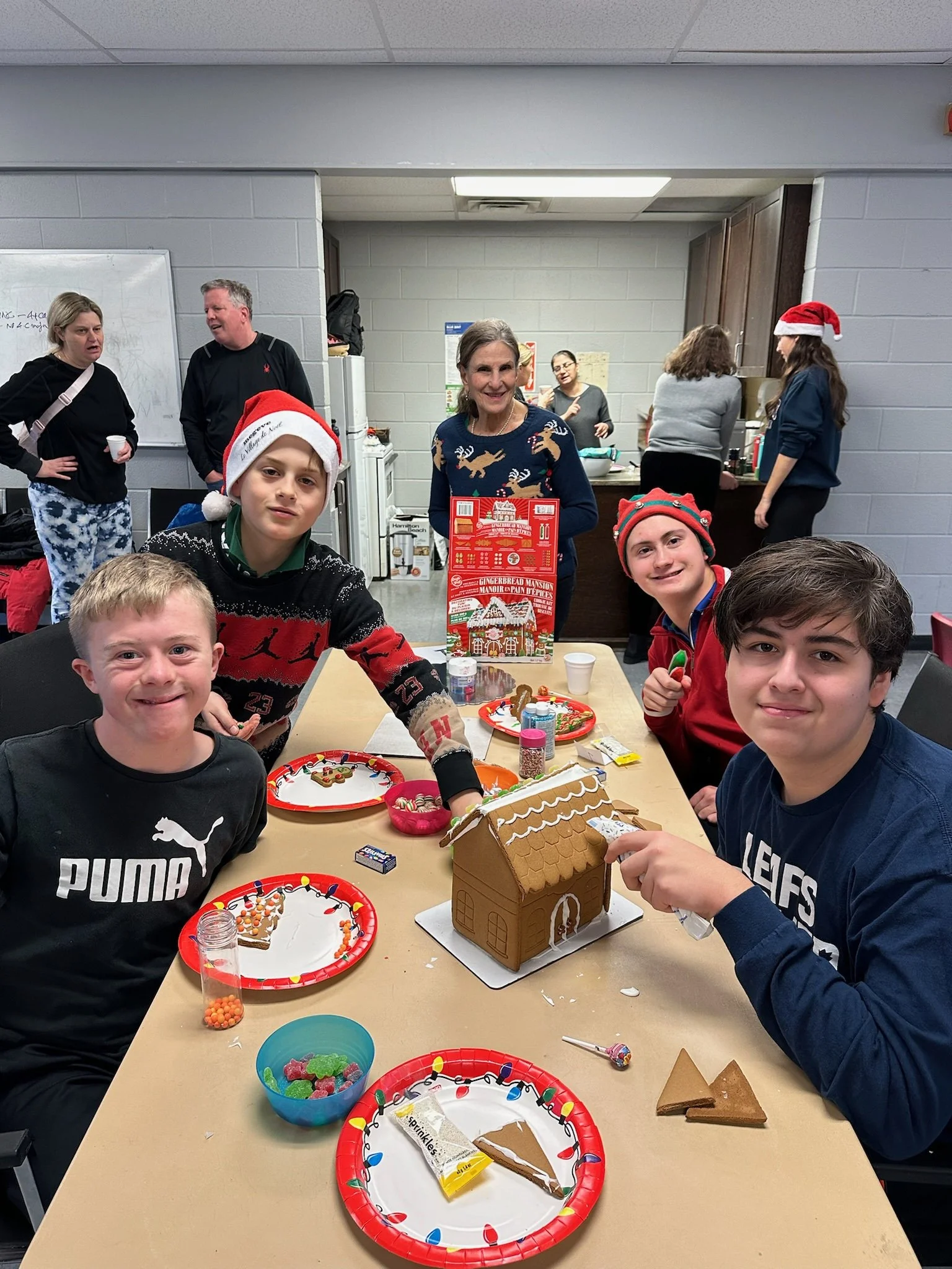 Children at a holiday party decorating a gingerbread house, with Christmas hats and a woman in the background wearing a festive sweater, in an indoor setting.