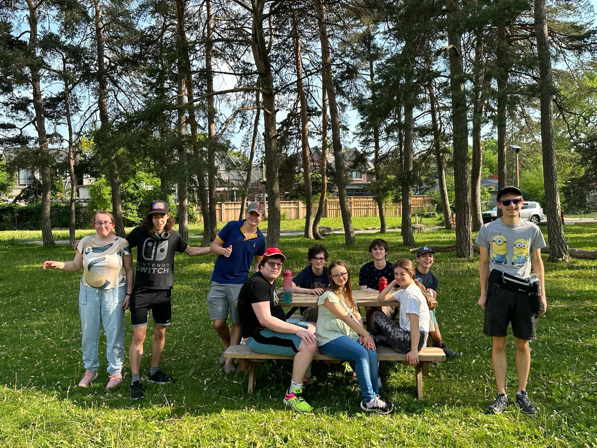 A group of young people sitting and standing around a picnic table in a park with trees and houses in the background.