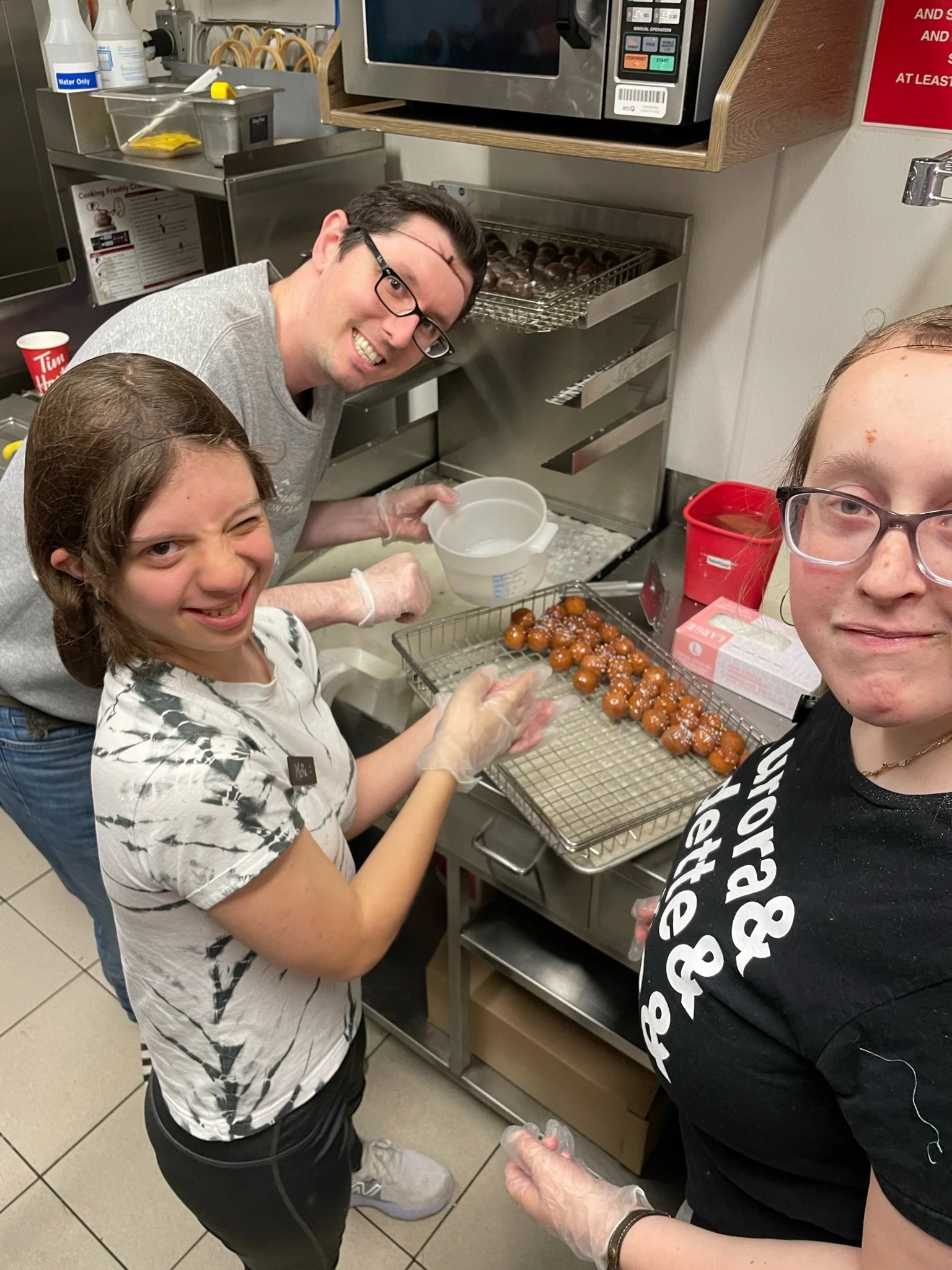 Three people in a kitchen are decorating and preparing doughnut holes with white icing, standing near a tray of ready doughnuts on a cooling rack.