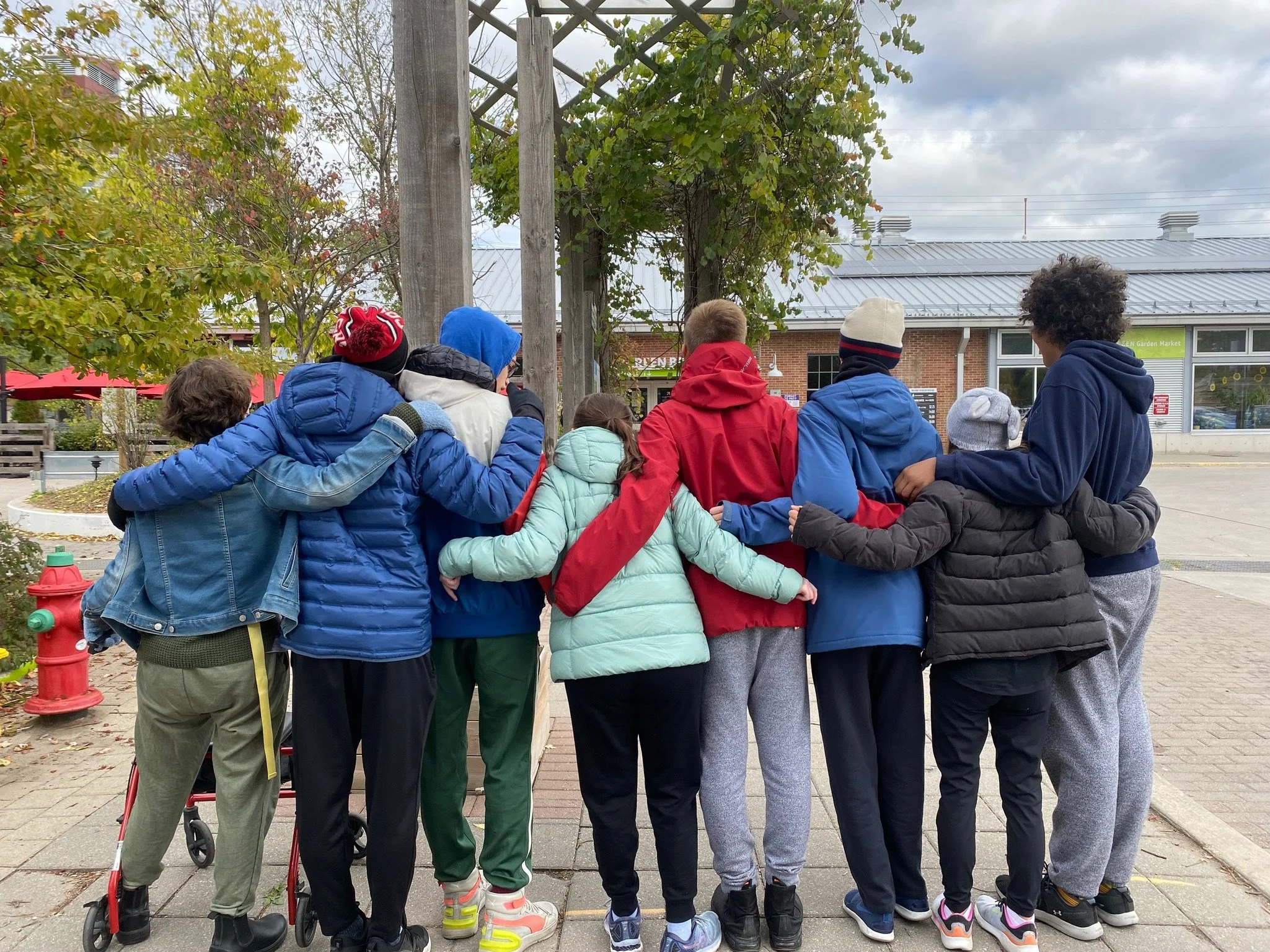 A group of children standing outdoors with their arms around each other, facing away from the camera.