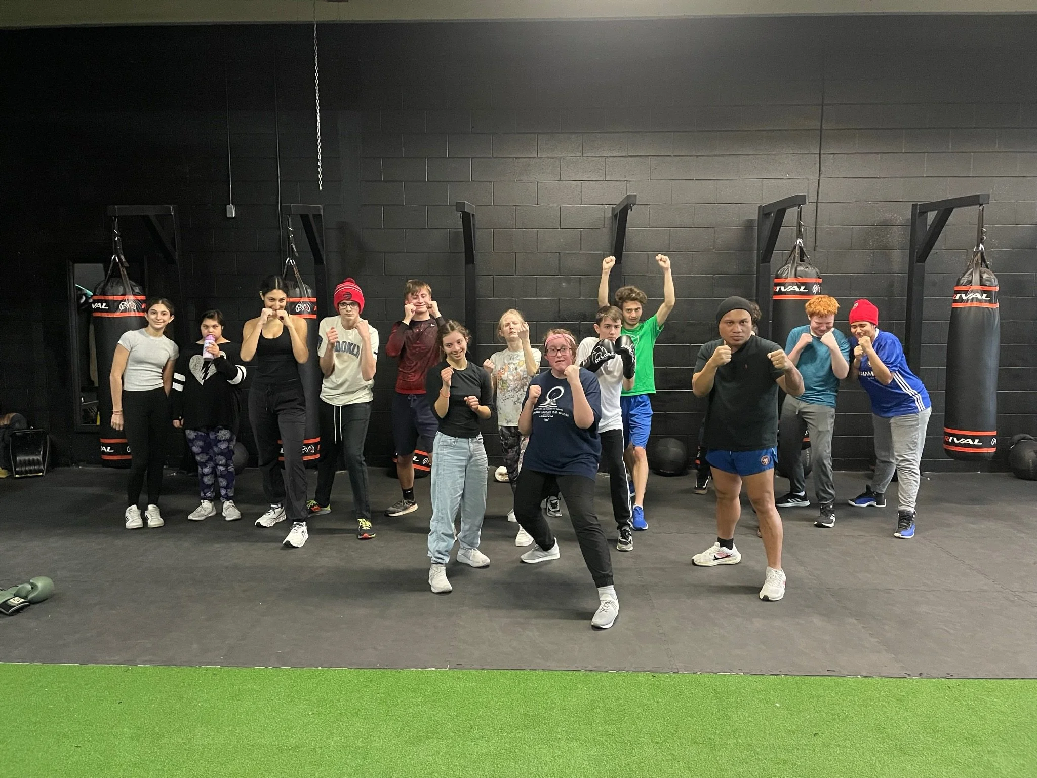 Group of people in a boxing gym posing in fighting stances, some with fists raised, in front of black punching bags on a dark wall, with some green artificial turf on the floor.