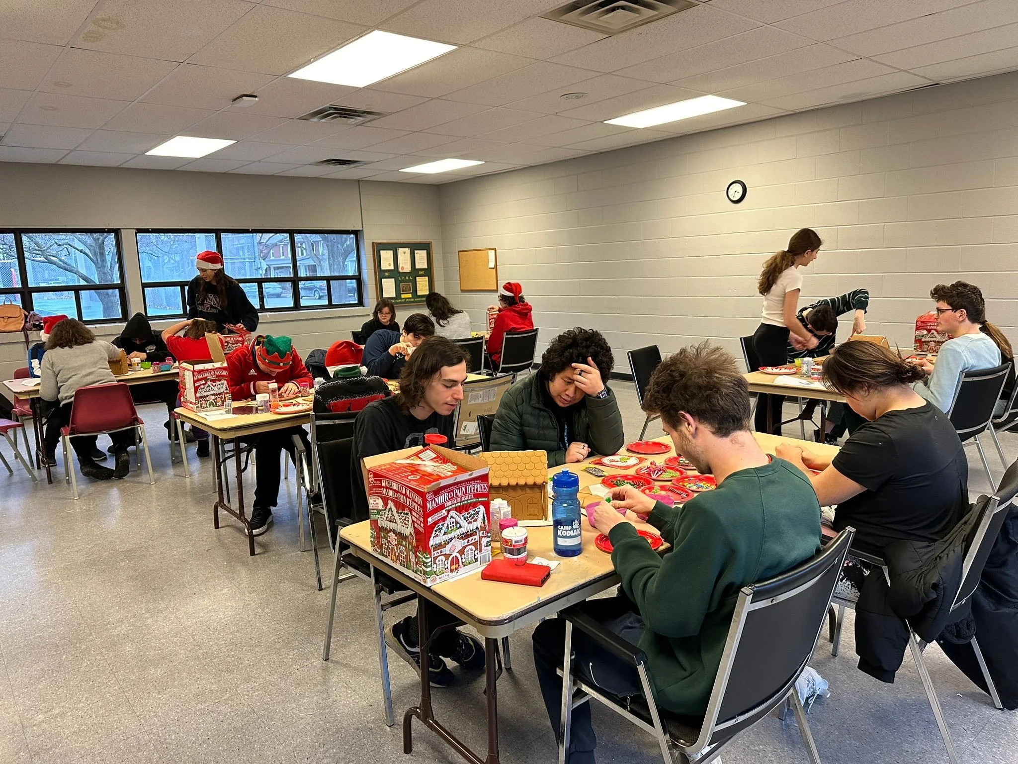 People gathered in a room decorating holiday cookies, with some wearing Santa hats, suggesting a Christmas celebration or party.