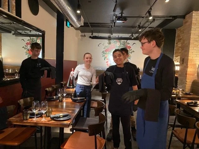 Four people in a restaurant, two men and two women, one woman giving a thumbs-up, the other woman smiling, the man in an apron talking to the group, tables set with glasses and plates.