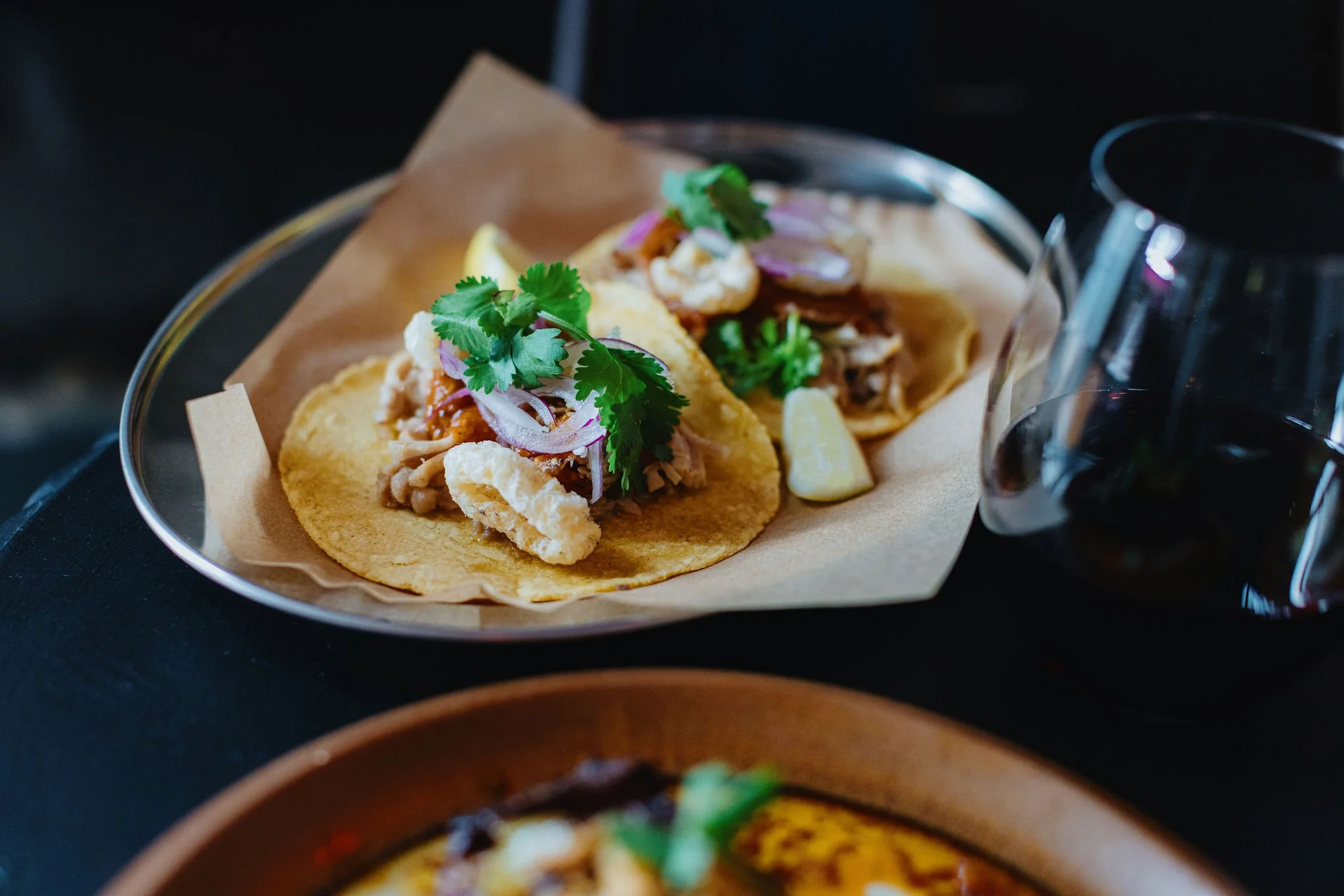 Two tacos on a plate with cilantro, onions, and lime wedge, alongside two glasses of red wine.