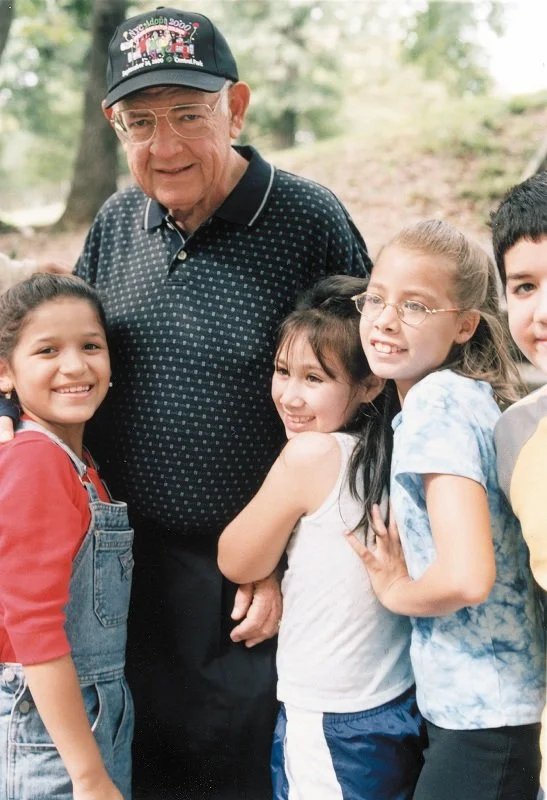 Older man in a black cap and glasses surrounded by four smiling children outdoors.