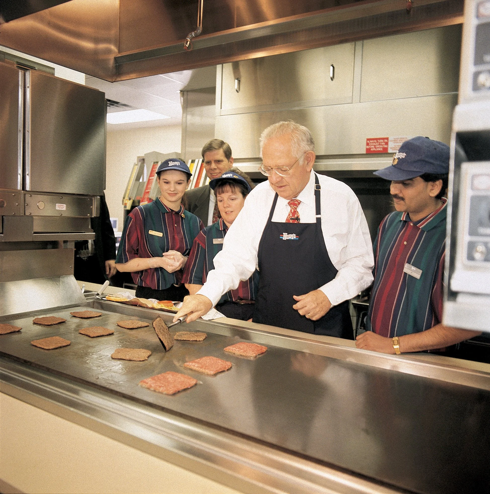 Employees in a kitchen preparing hamburgers, with hamburger patties on a grill in the foreground.