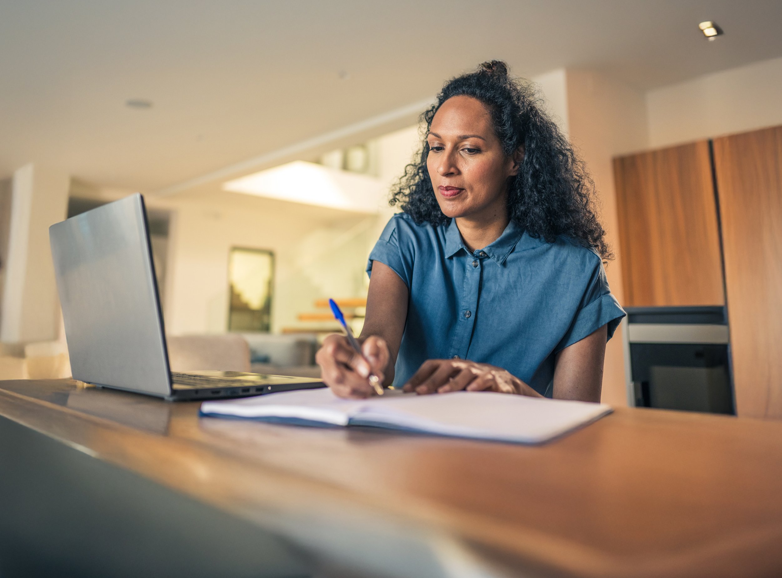 Woman sitting at a table, writing in a notebook with a pen, with a laptop open beside her in a bright, modern kitchen or office space.