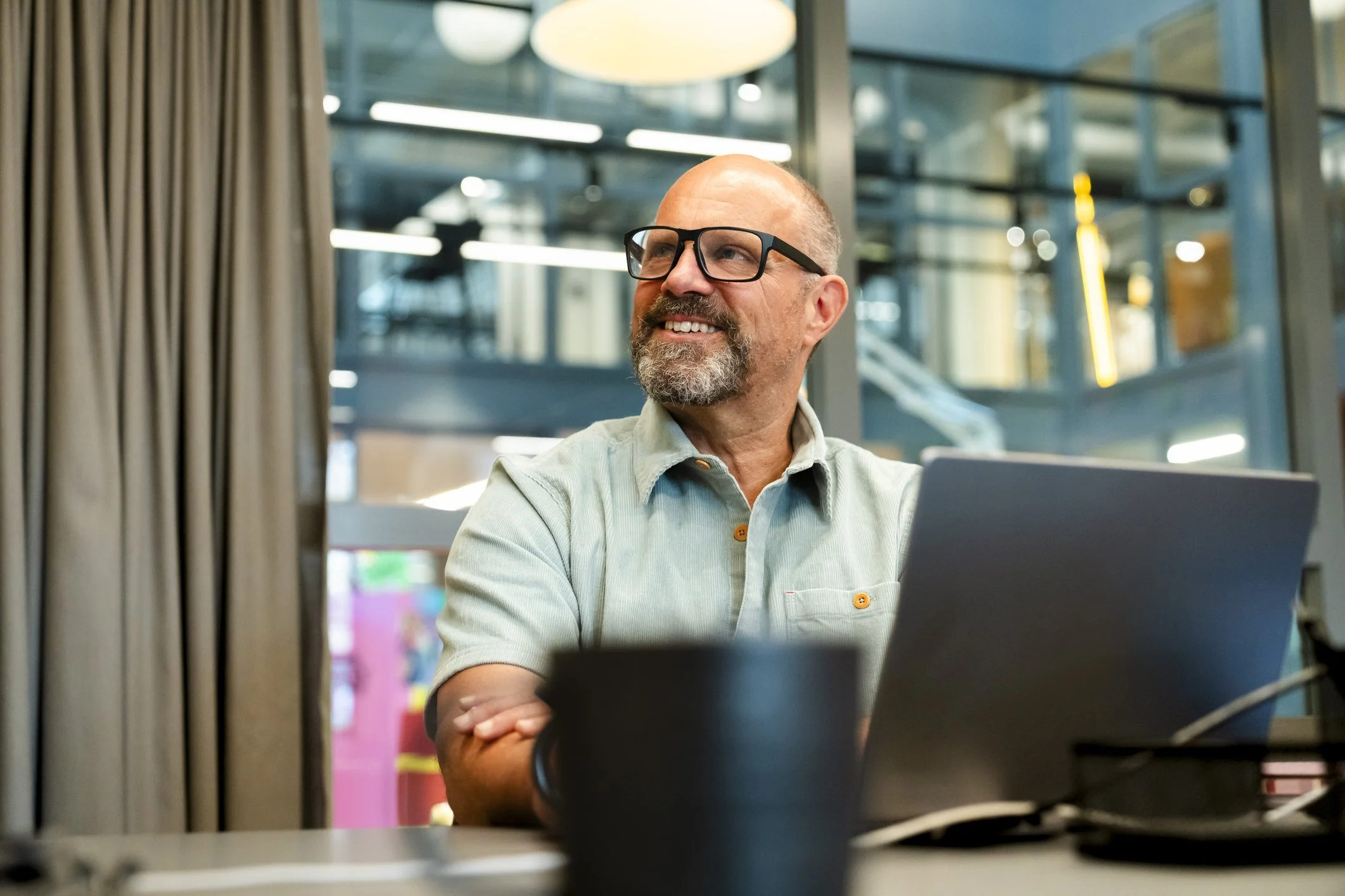 A middle-aged man with glasses and a beard, smiling and sitting at a desk with a laptop and coffee mug, in a modern office or coworking space with large windows and industrial decor.