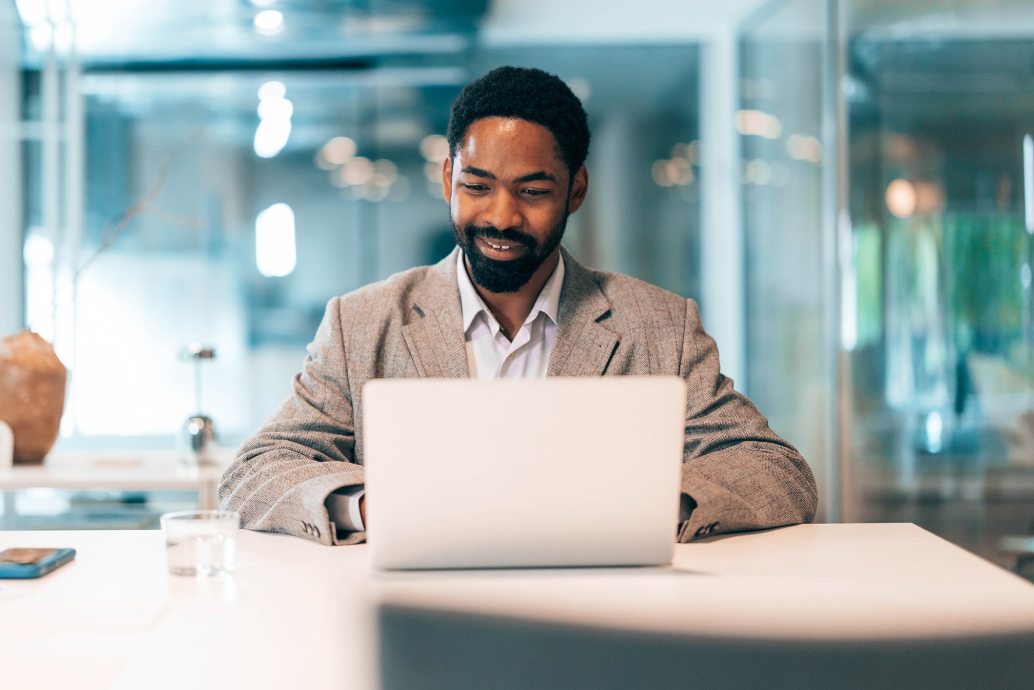 A man in a grey blazer working on a laptop at a white desk in a modern office setting.